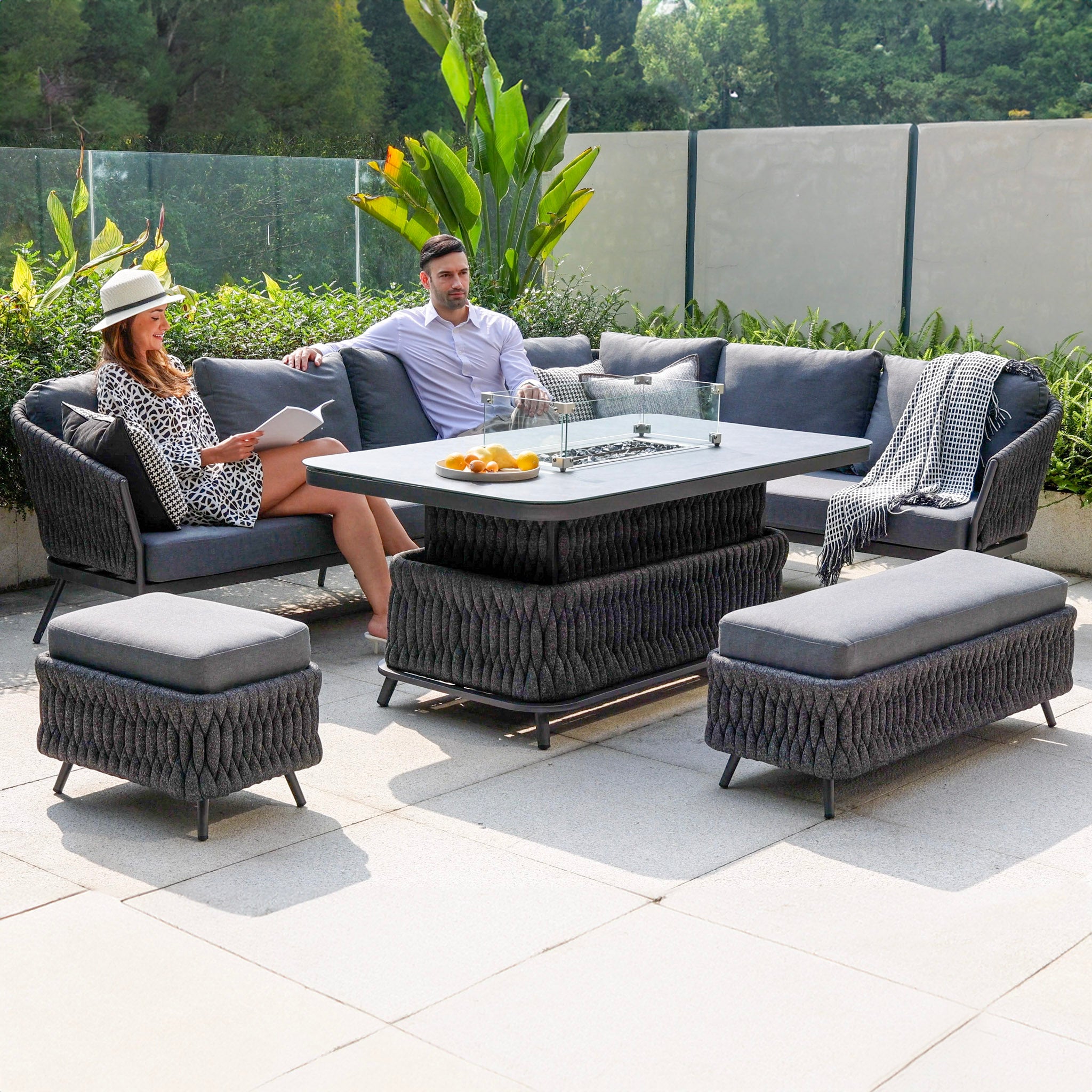 Two people relax on the Palma Rope Rectangular Corner Dining Set with Rising Firepit Table in Grey (Right Hand), featuring a water-repelling thick rope weave and sturdy aluminium frames, surrounded by greenery, enjoying a sunny day.