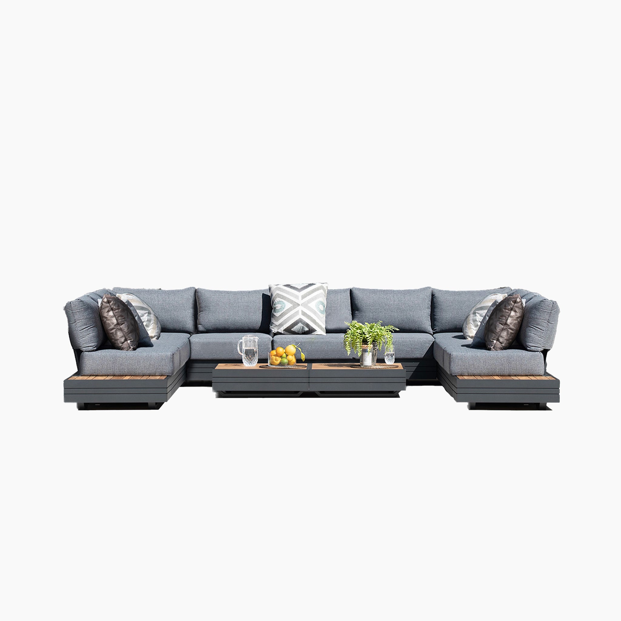The Panama Luxury Outdoor Large U-Shape Corner Sofa Set in Washed Grey features a stunning gray modular sectional sofa adorned with patterned throw pillows. Pair it with a central table displaying fruit and drinks, and add a potted plant for an inviting setup. This exquisite piece is part of the Harbour Lifestyle Panama collection, crafted from sustainable teak to offer an eco-friendly touch of elegance.