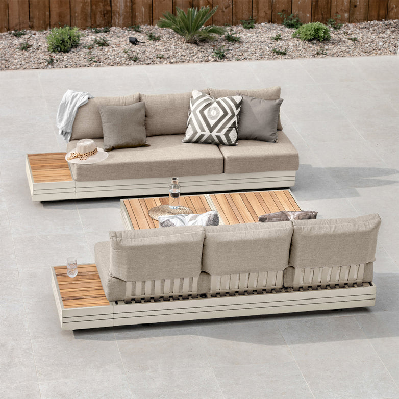 Two modern Panama 3 Seat Sofas with Sun Lounger Feature in Latte, featuring beige cushions and wooden accents, are arranged on a patio next to a wooden table.