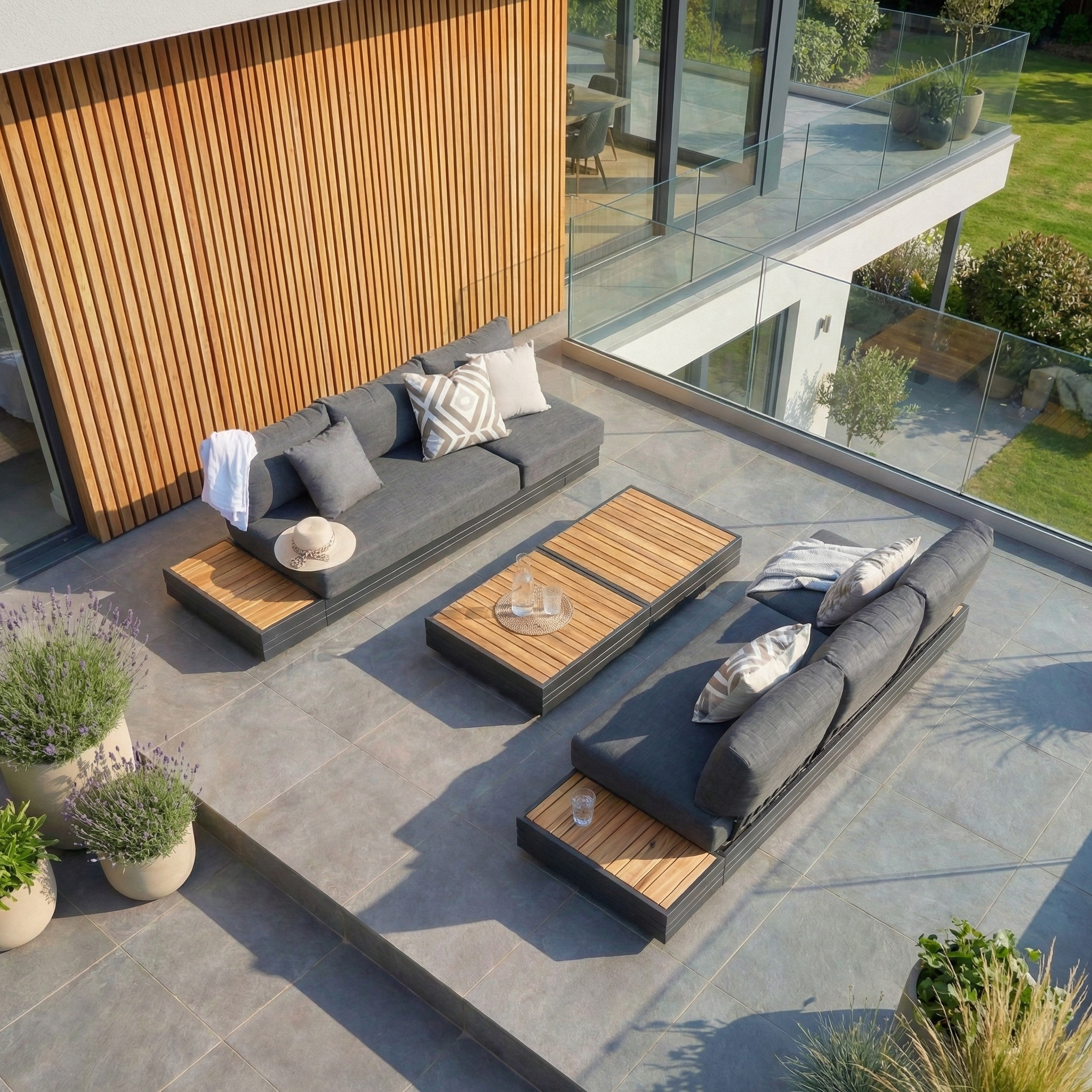 Modern outdoor patio featuring a Panama 3 Seat Sofa with Sun Lounger Feature in Charcoal, wooden tables, potted plants, and glass railing overlooking a green lawn.