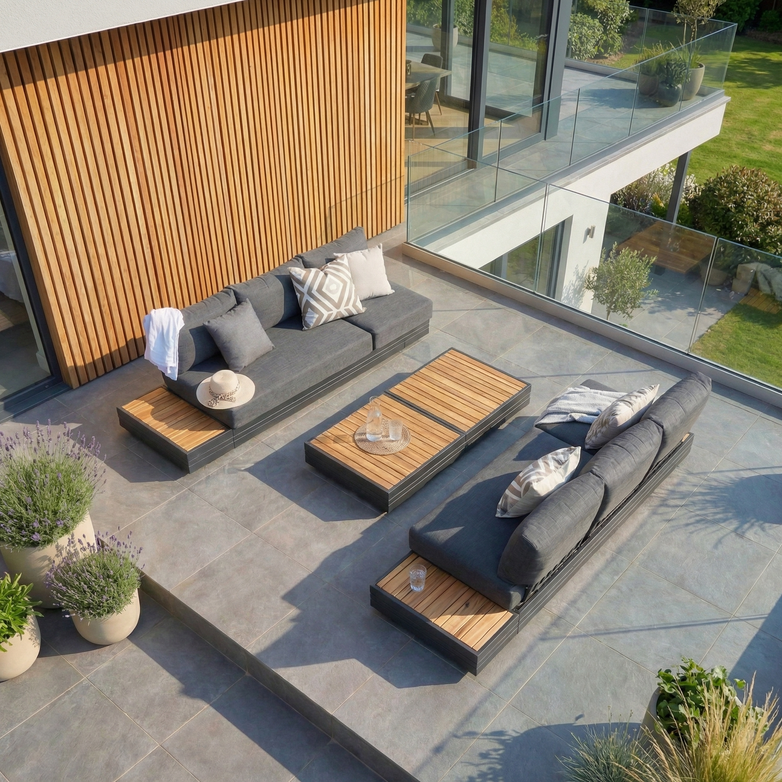 Modern outdoor patio featuring a Panama 3 Seat Sofa with Sun Lounger Feature in Charcoal, wooden tables, potted plants, and glass railing overlooking a green lawn.