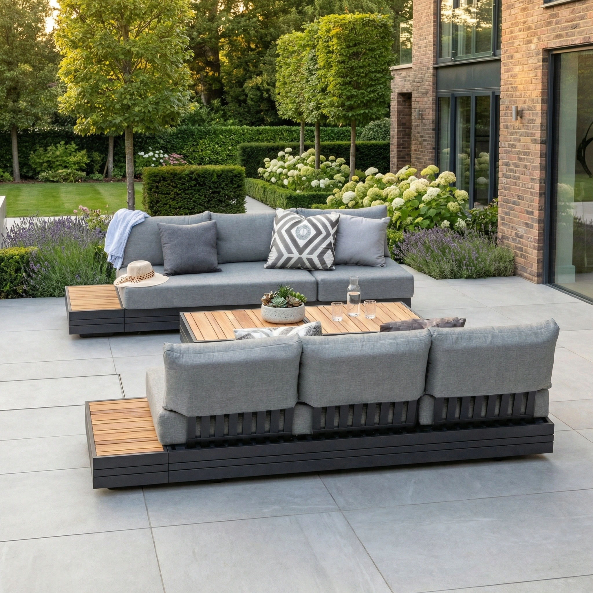 Modern patio with the Panama 3 Seat Sofa featuring a sun lounger in washed grey, complemented by wooden tables, potted plants, and a lush green garden—the ideal spot to unwind with stylish outdoor furniture.