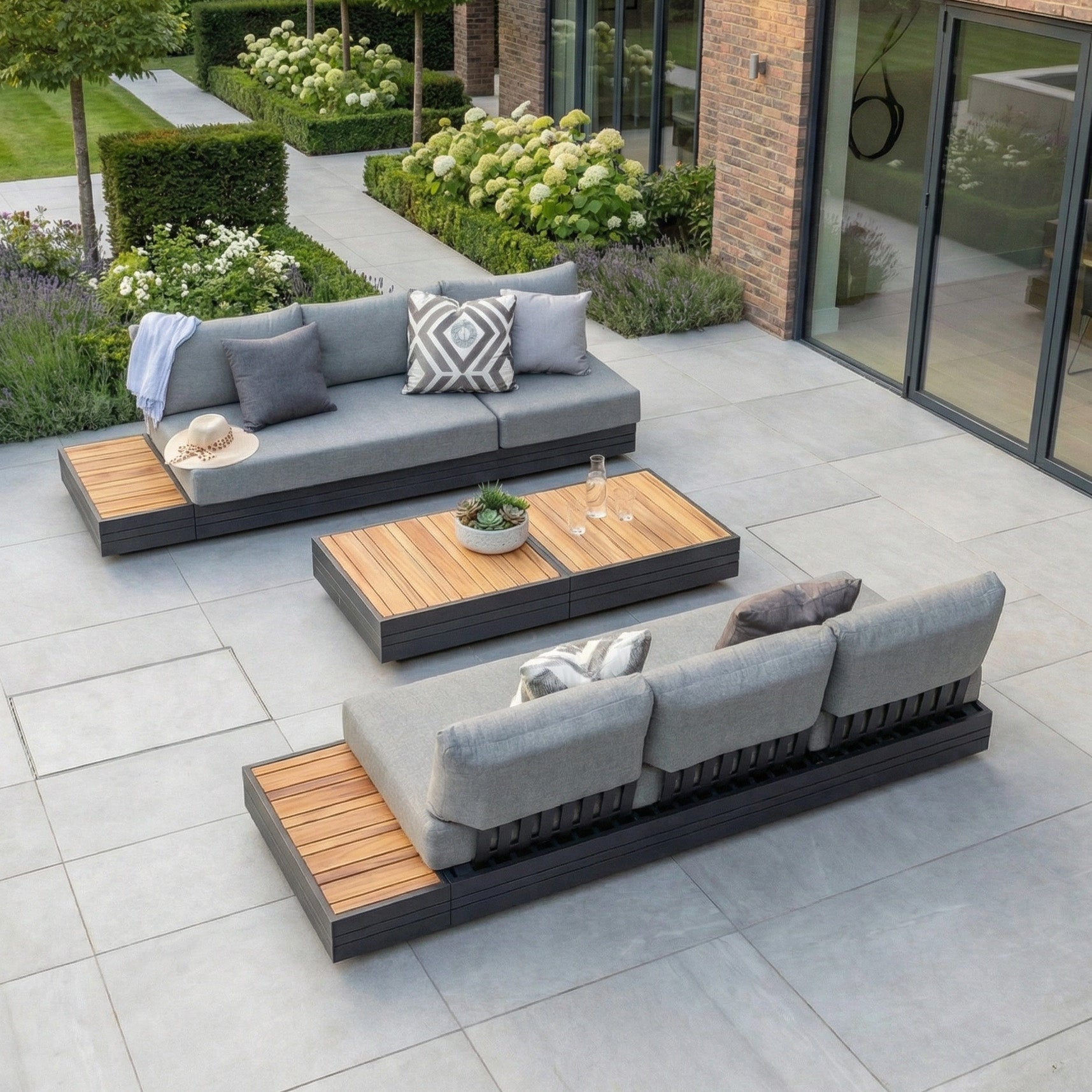 Contemporary outdoor patio featuring the Panama 3 Seat Sofa with Sun Lounger Feature in Washed Grey, complemented by wooden tables, gray cushions, and lush greenery—a perfect spot for relaxing on stylish garden furniture.
