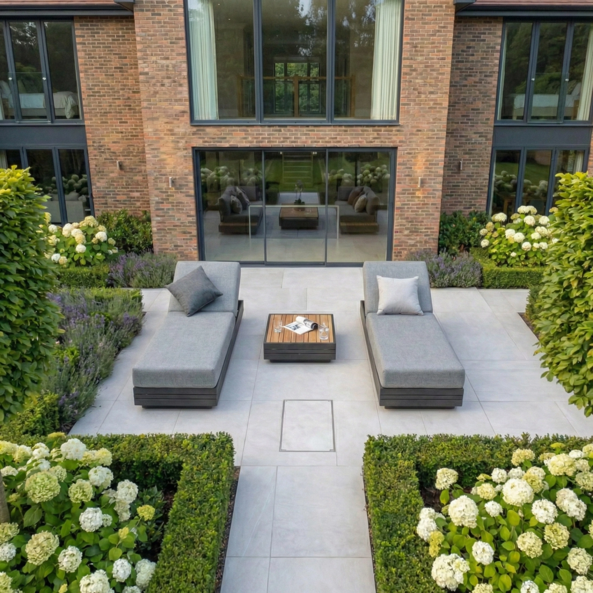 A Panama 3 Seat Sofa with Sun Lounger Feature in Washed Grey sits on a patio bordered by trimmed hedges and white flowers, creating an inviting outdoor furniture setting.