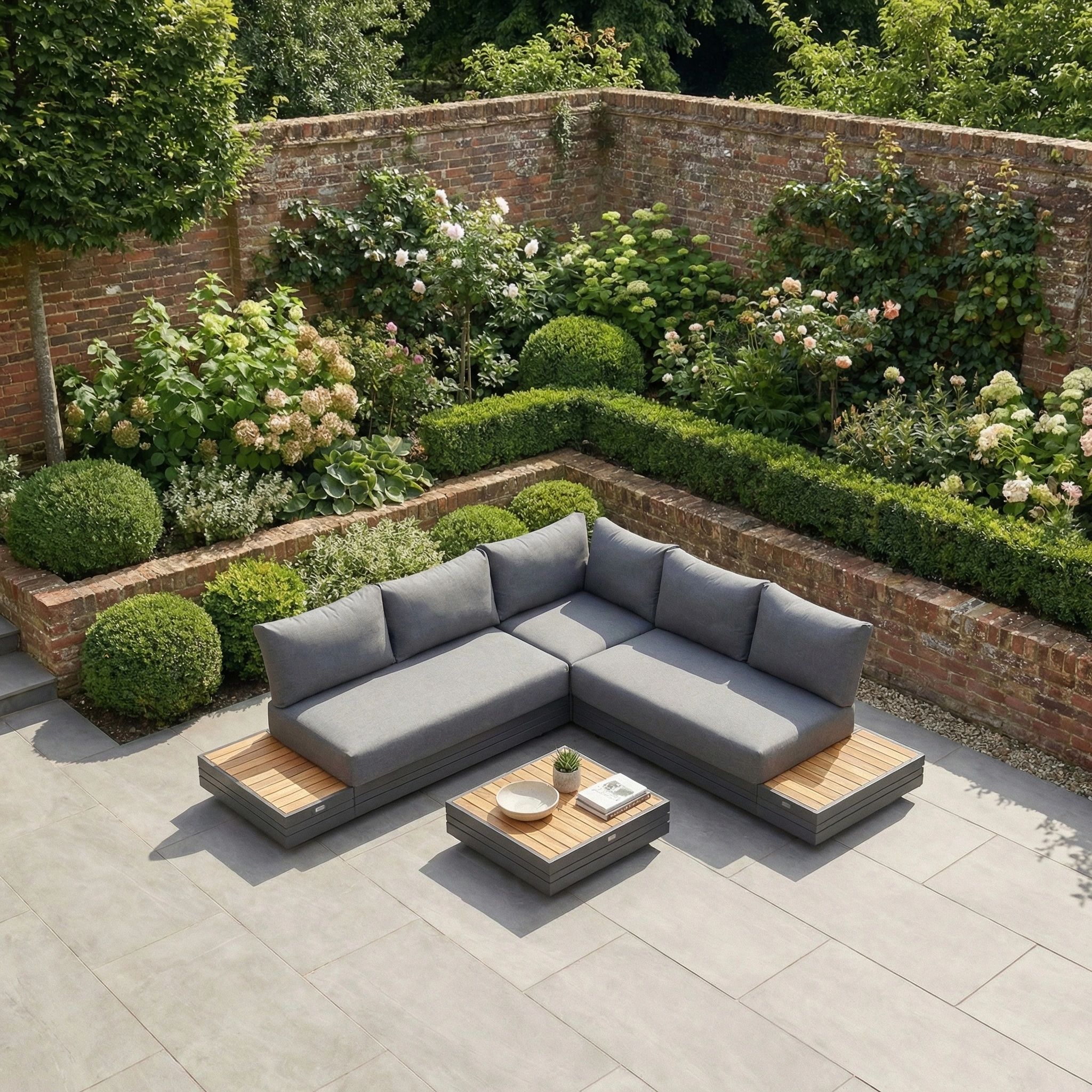Panama Luxury Outdoor Corner Group Set in Charcoal with a modular sofa and wooden side tables, displayed on a stone patio amid lush greenery and a brick wall.
