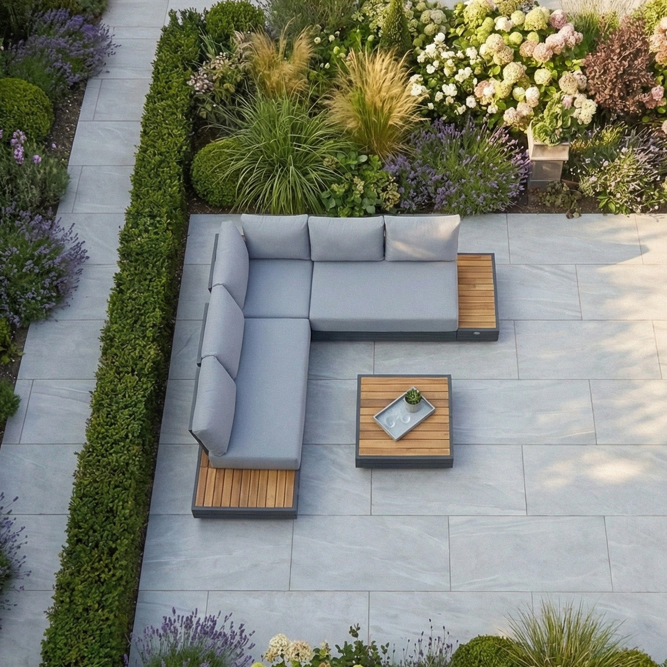 The Panama Luxury Outdoor Corner Group Set in Washed Grey, with its modern L-shaped design and table, creates a stylish outdoor living space on any stone patio, beautifully complemented by lush greenery and flowering plants.