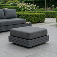 The Panama Corner Unit/Footstool in Charcoal with a gray cushion sits on a tiled patio beside a matching Panama sofa and lush green hedges.