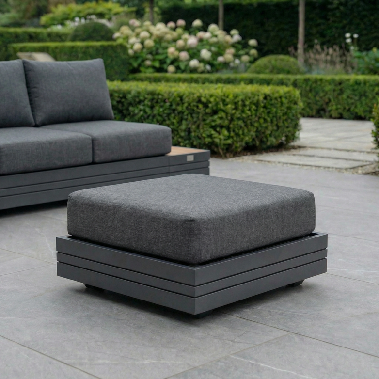 The Panama Corner Unit/Footstool in Charcoal with a gray cushion sits on a tiled patio beside a matching Panama sofa and lush green hedges.