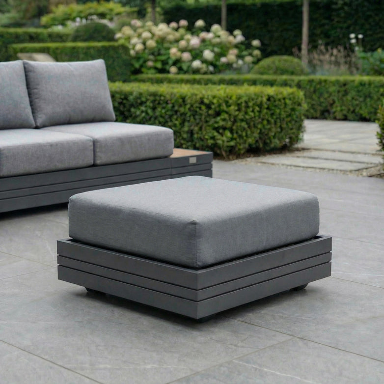 The Panama Corner Unit/Footstool in Washed Grey, with gray cushions, is arranged on a tiled terrace surrounded by lush hedges and greenery, creating a chic outdoor seating area.