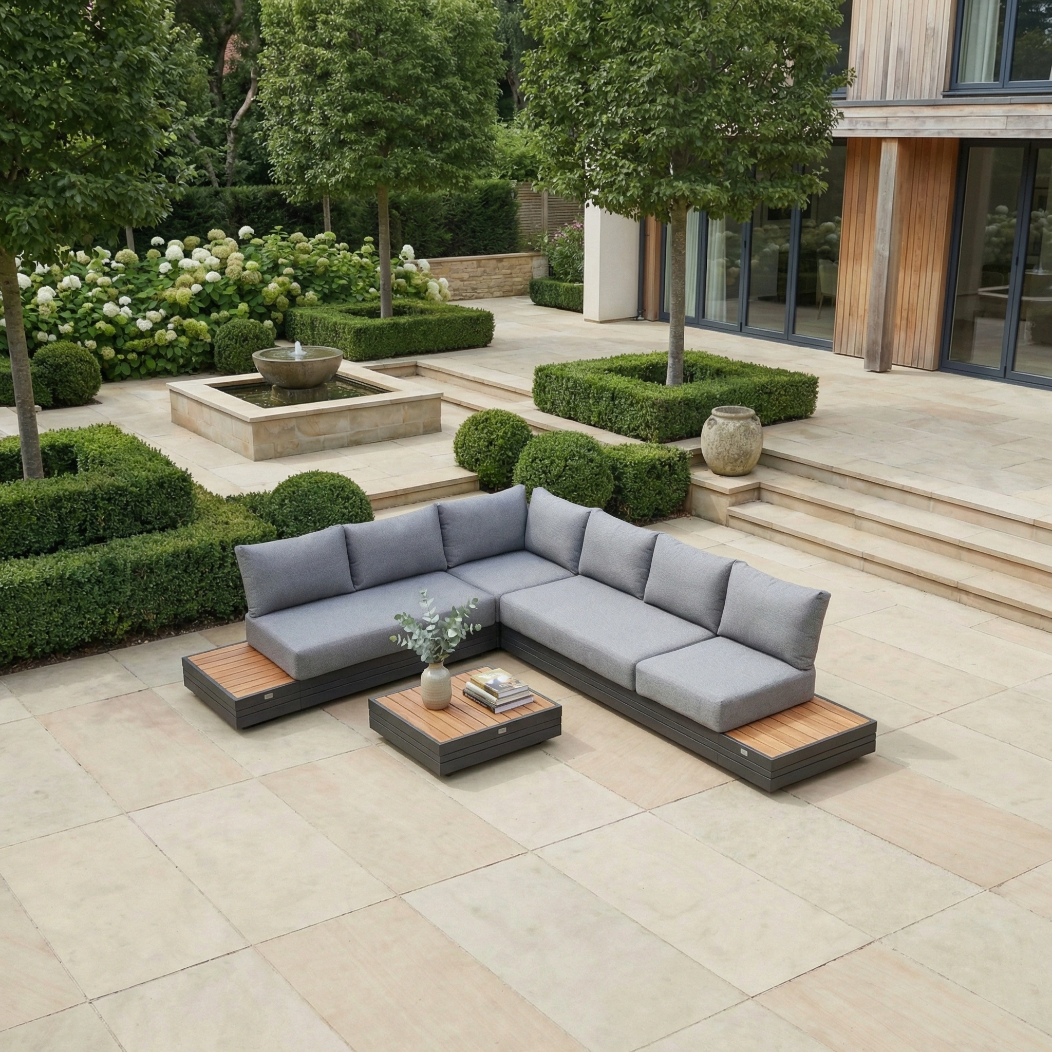 The Panama Luxury Outdoor L-Shape Corner Group Set in Washed Grey, paired with a coffee table, sits on a stone patio surrounded by trimmed greenery and a tiered fountain—perfect for stylish outdoor relaxation.