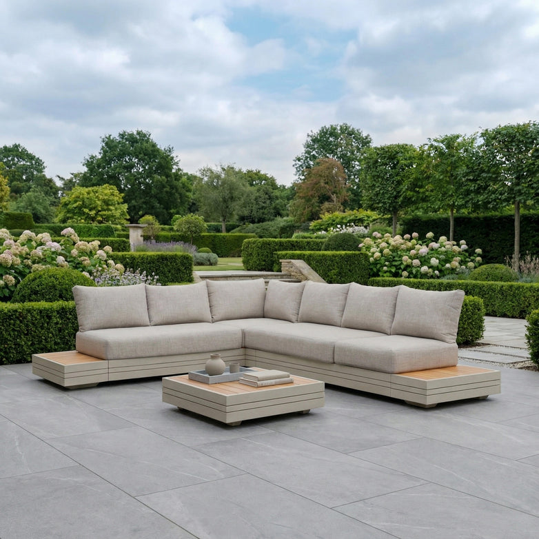 The Panama Luxury Outdoor L-Shape Corner Group Set in Latte with a coffee table sits on a spacious stone patio in a landscaped garden, expertly crafted from sustainable teak and durable aluminium.
