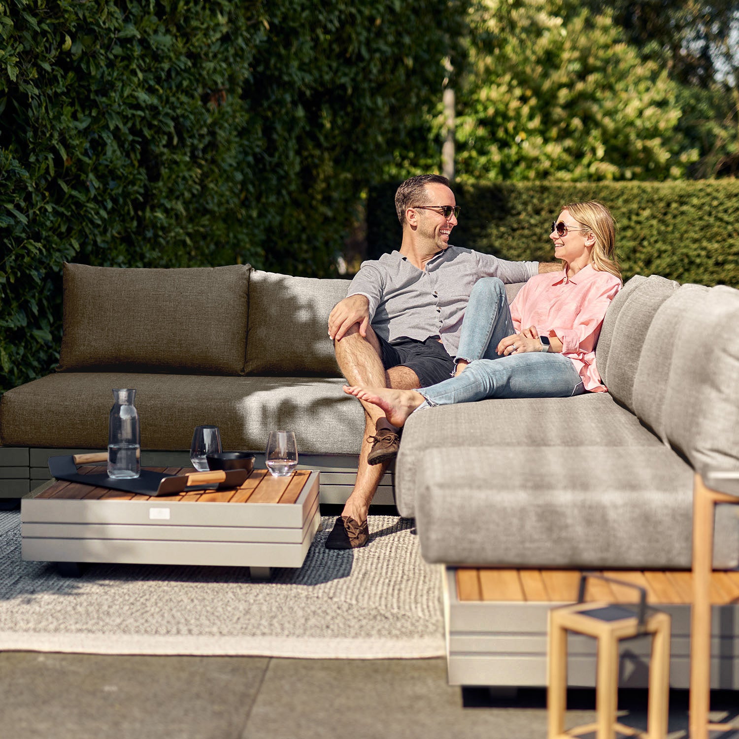 A couple sits on the Panama Luxury Outdoor L-Shape Corner Group Set in Latte, smiling at each other. A coffee table and a lush green hedge provide the perfect backdrop to their sustainable teak outdoor setting.