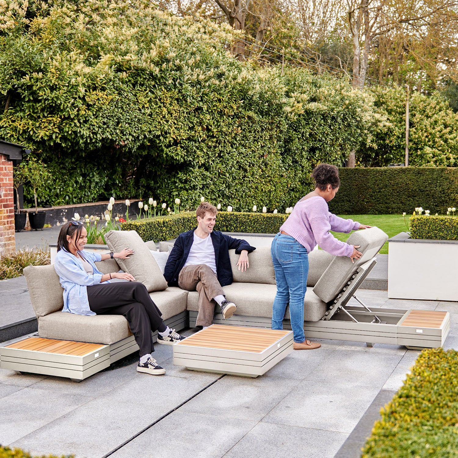 Three people relax on the Panama Luxury Outdoor L-Shape Corner Group Set in Latte from Harbour Lifestyle, situated in a garden. One woman is adjusting a section of the luxury outdoor set made of sustainable teak.