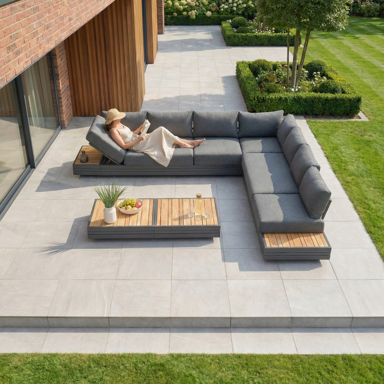 A woman relaxes with a book on the Panama Luxury Outdoor Large Corner Group Set in Charcoal, a modern modular garden set featuring sustainable teak tables on a tiled terrace beside a green lawn.