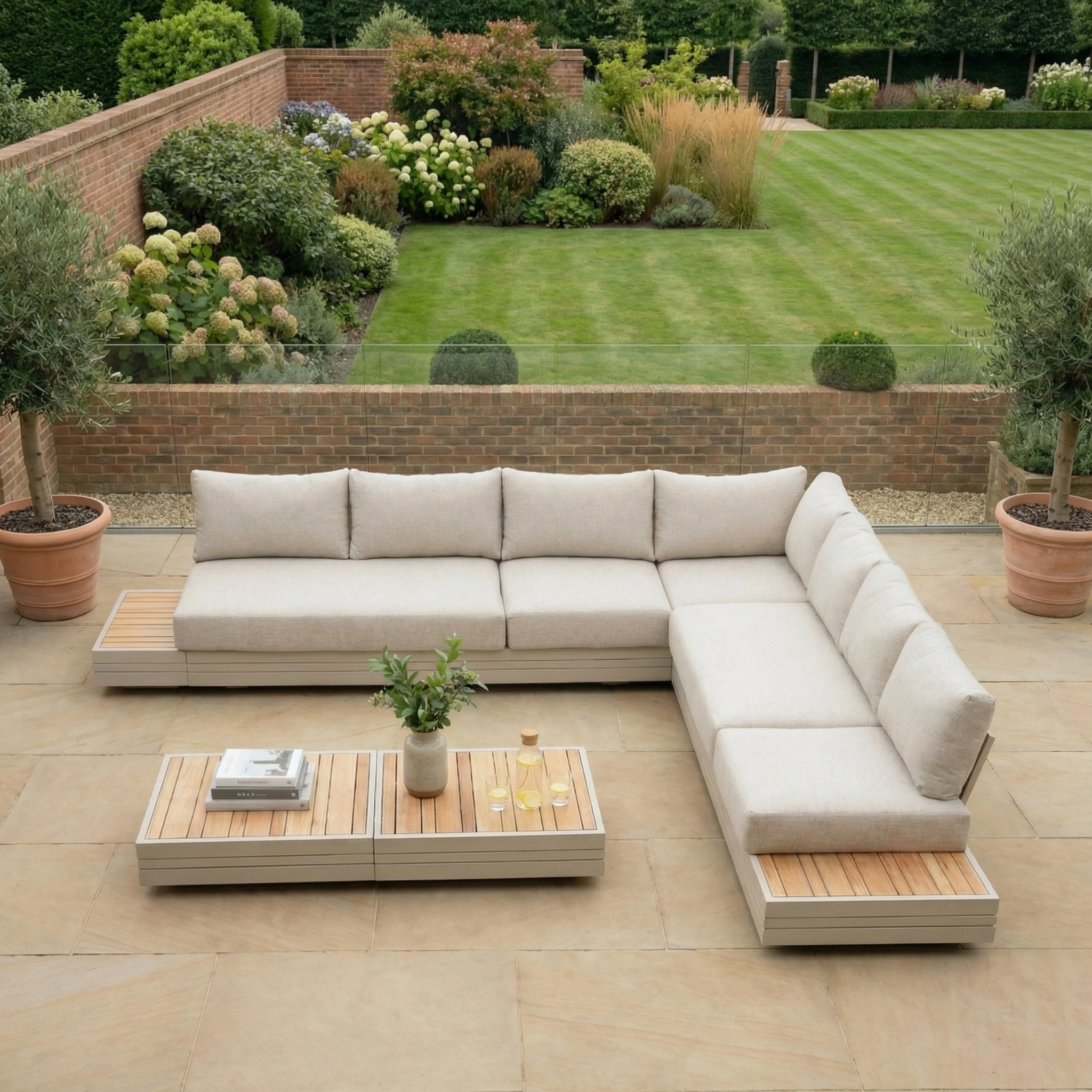 The Panama Luxury Outdoor Large Corner Group Set in Latte with low tables on a stone patio, overlooking a landscaped garden, creates an inviting retreat with sustainable teak accents.