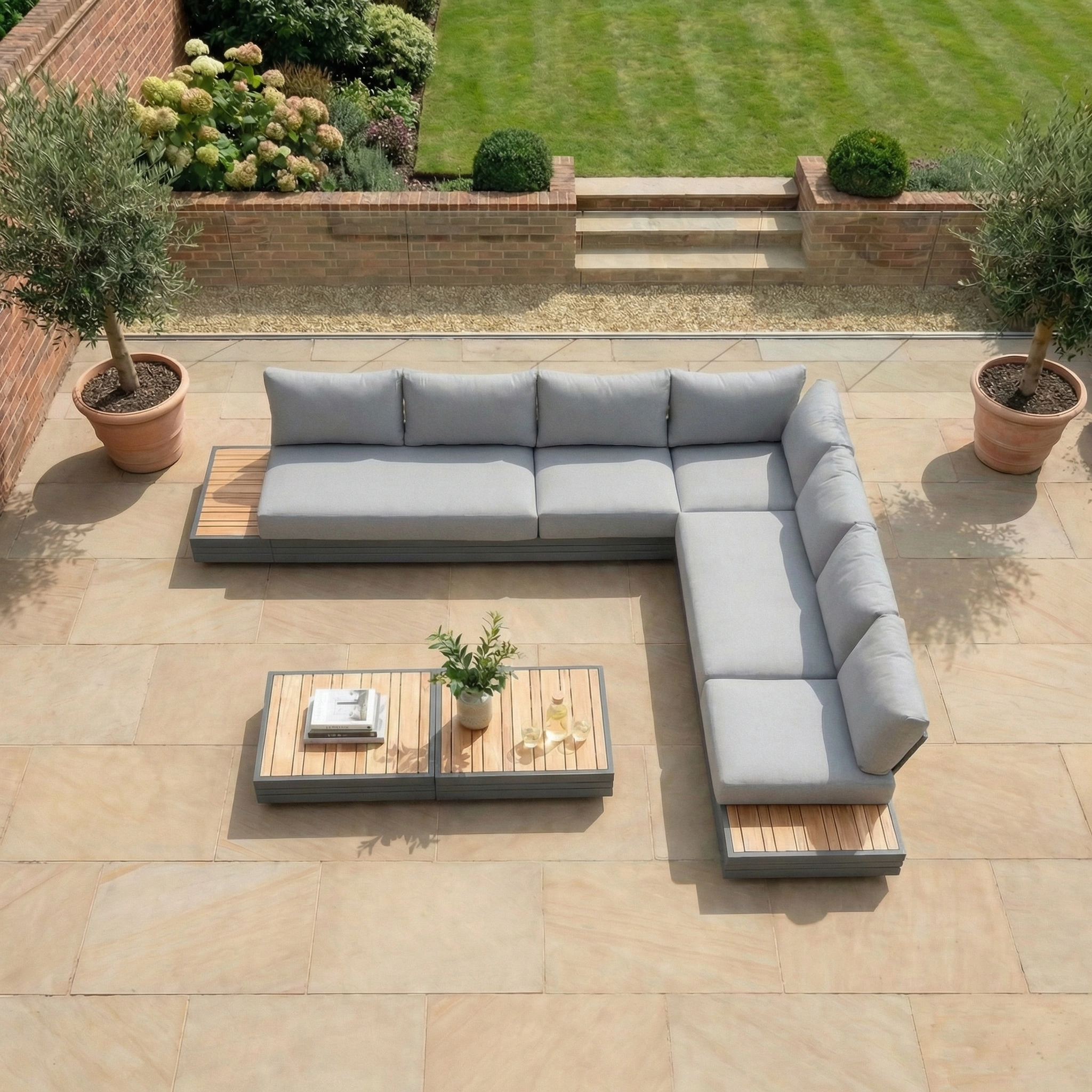 The Panama Luxury Outdoor Large Corner Group Set in Washed Grey is arranged on a stone patio with coffee tables, overlooking a manicured lawn and garden.