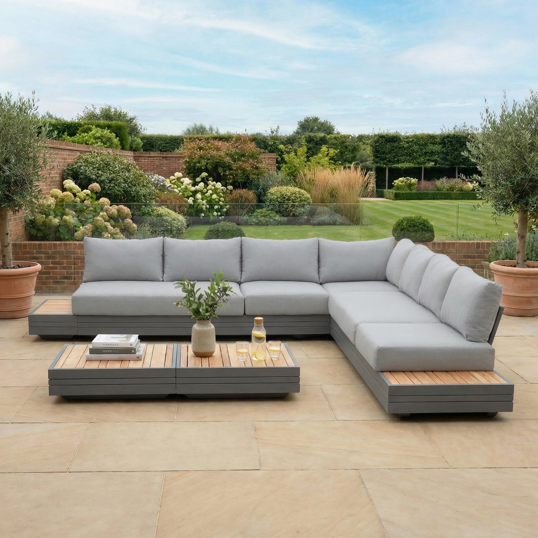 The Panama Luxury Outdoor Large Corner Group Set in Washed Grey, paired with coffee tables, sits on a stone patio surrounded by lush garden greenery.