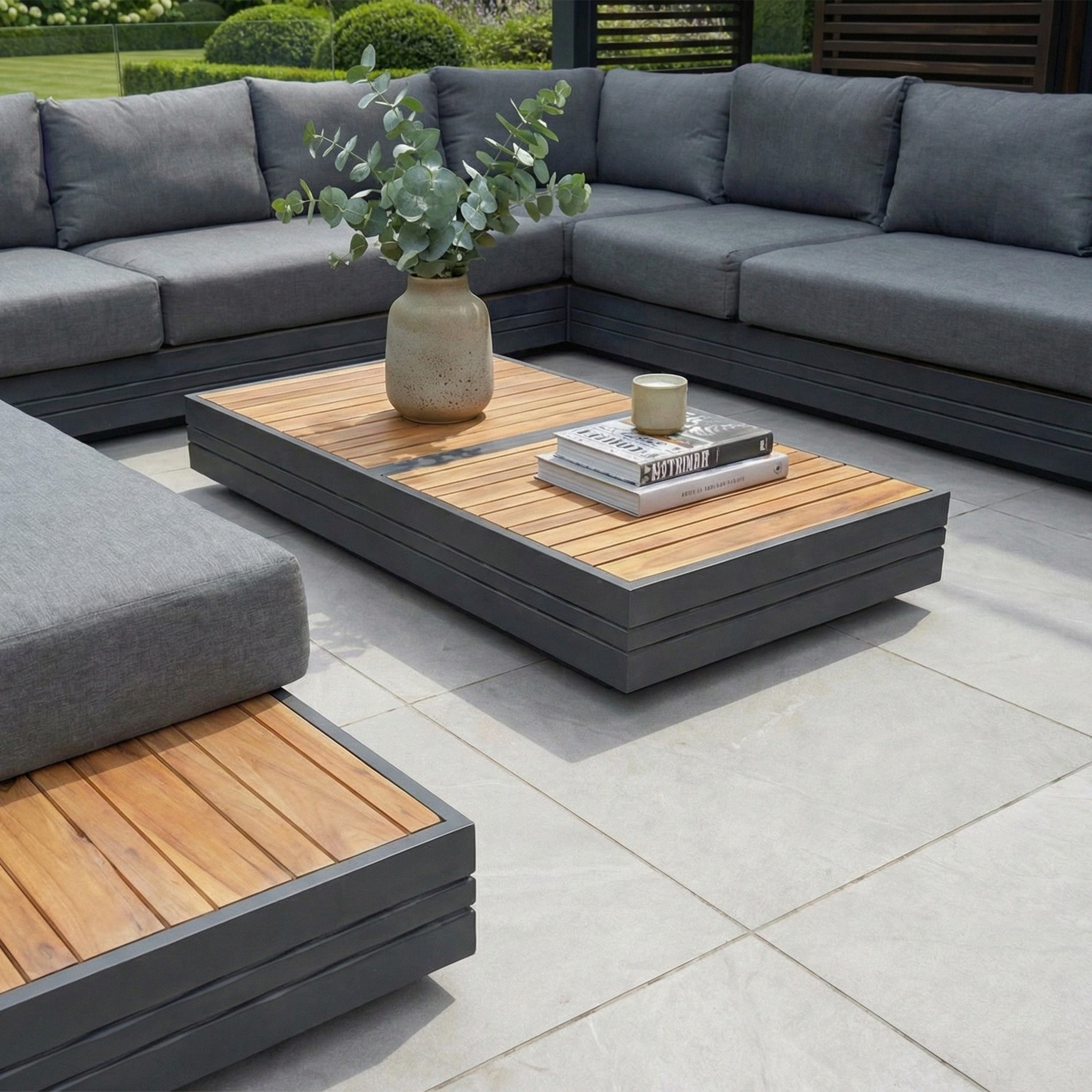 The Panama Luxury Outdoor Large U-Shape Corner Sofa Set in Charcoal features gray cushions, a sustainable teak wooden table, and is styled with greenery, books, and a candle on a tiled patio floor.