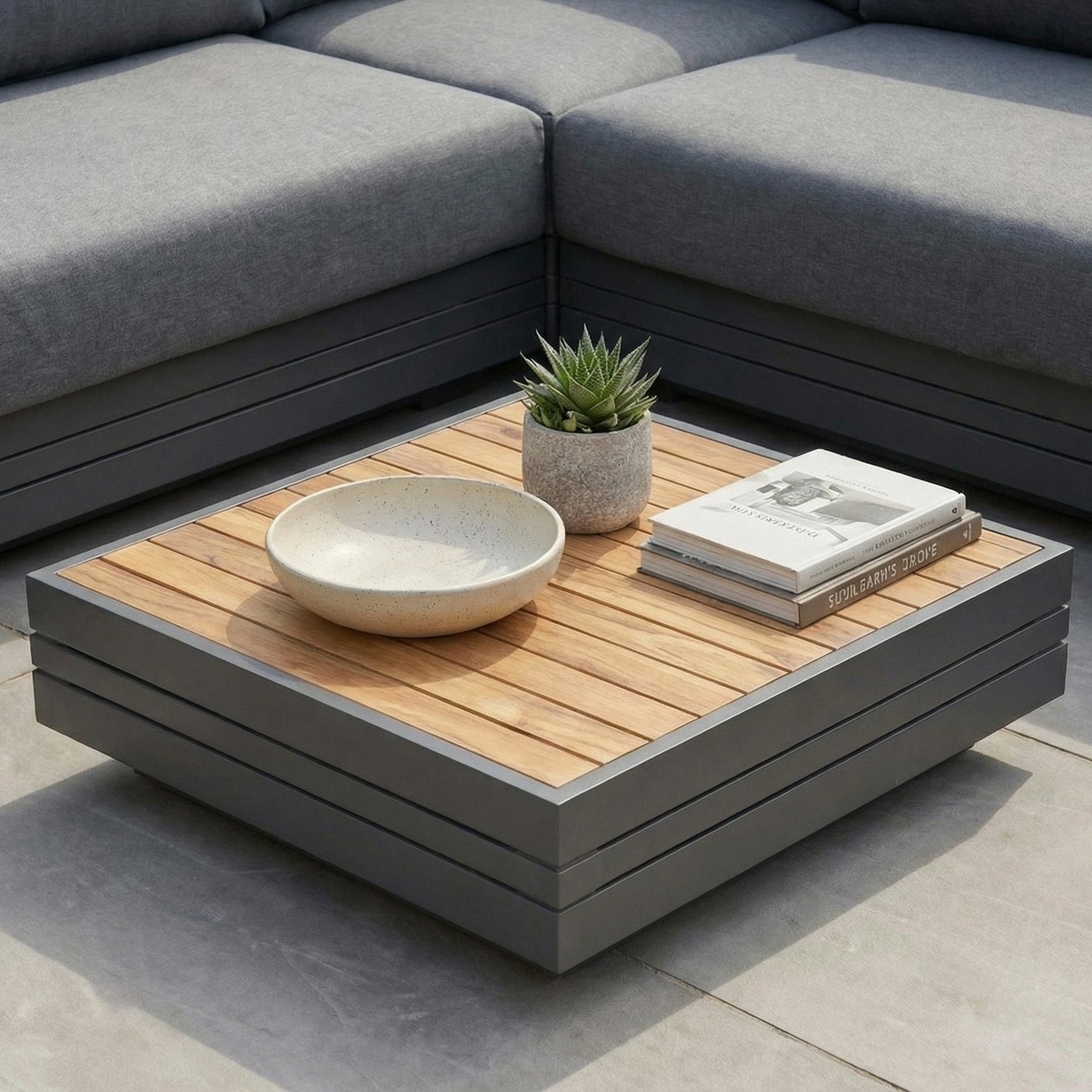 The Panama Teak Coffee Table, styled with a plant, bowl, and books beside a gray sectional on the patio, creates a perfect luxury outdoor coffee table setup.