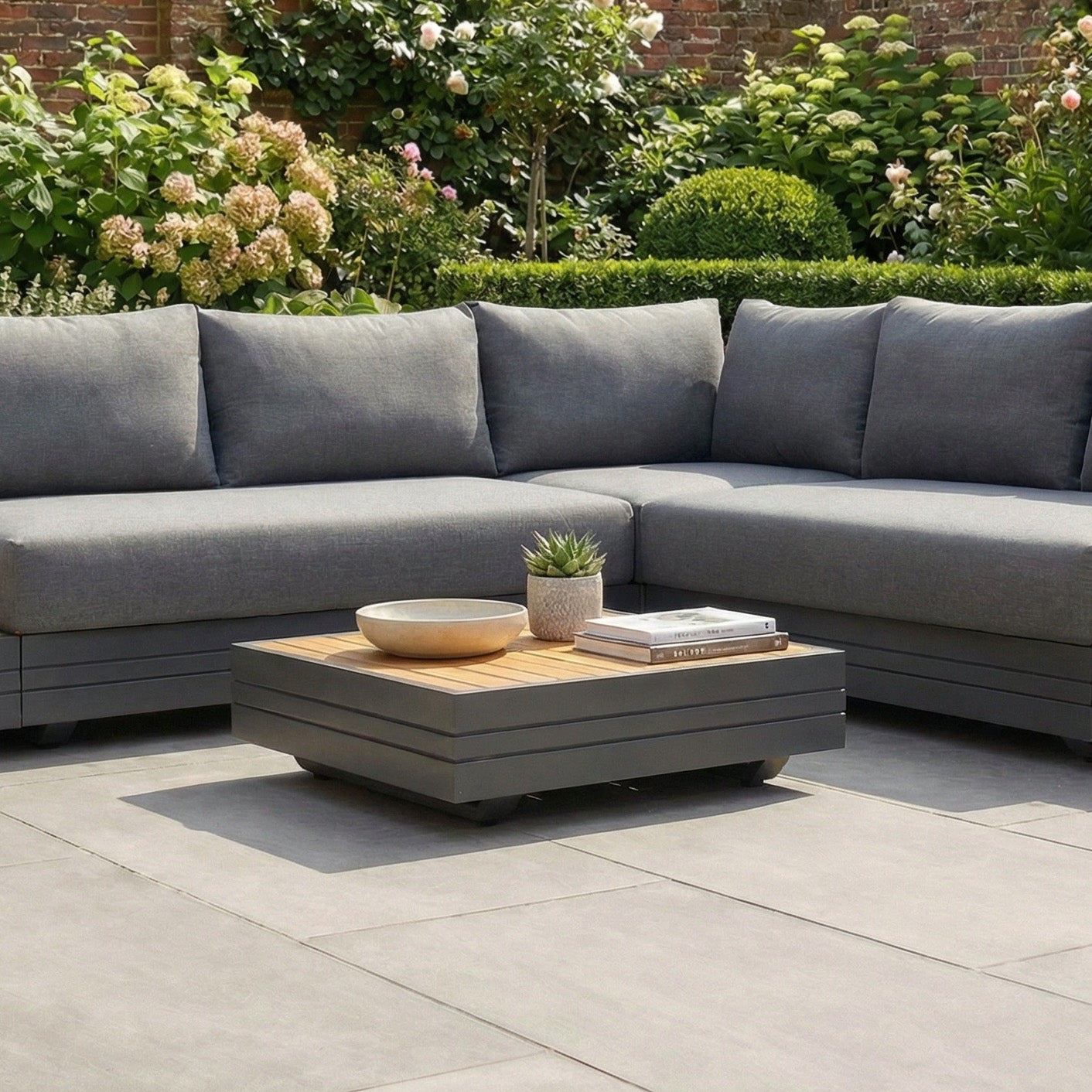 Gray outdoor sectional sofa paired with the Panama Teak Coffee Table, styled with books and a plant, set on a patio in a garden.