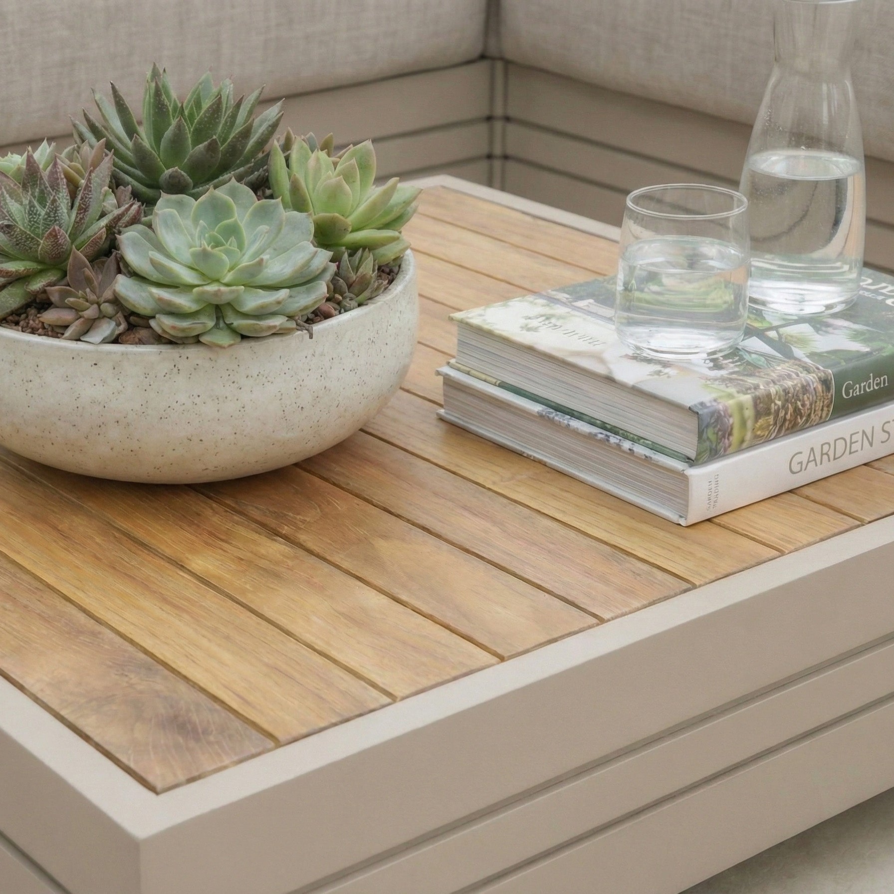 The Panama Teak Coffee Table in Latte, styled with potted succulents, two books, a glass of water, and a carafe, creates a charming outdoor coffee table scene.