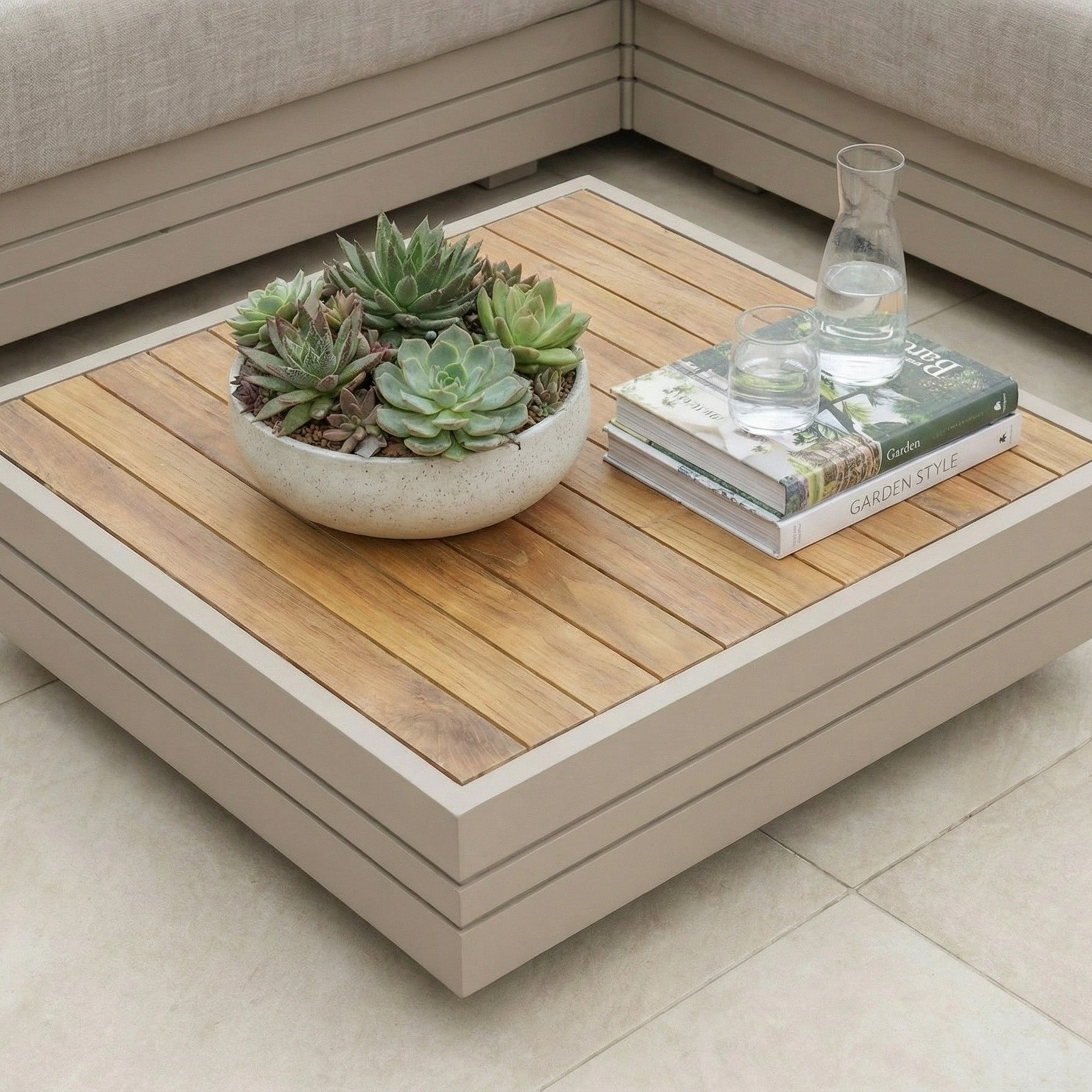 The Panama Teak Coffee Table in Latte is topped with a bowl of succulents, books, a glass carafe, and two glasses of water.