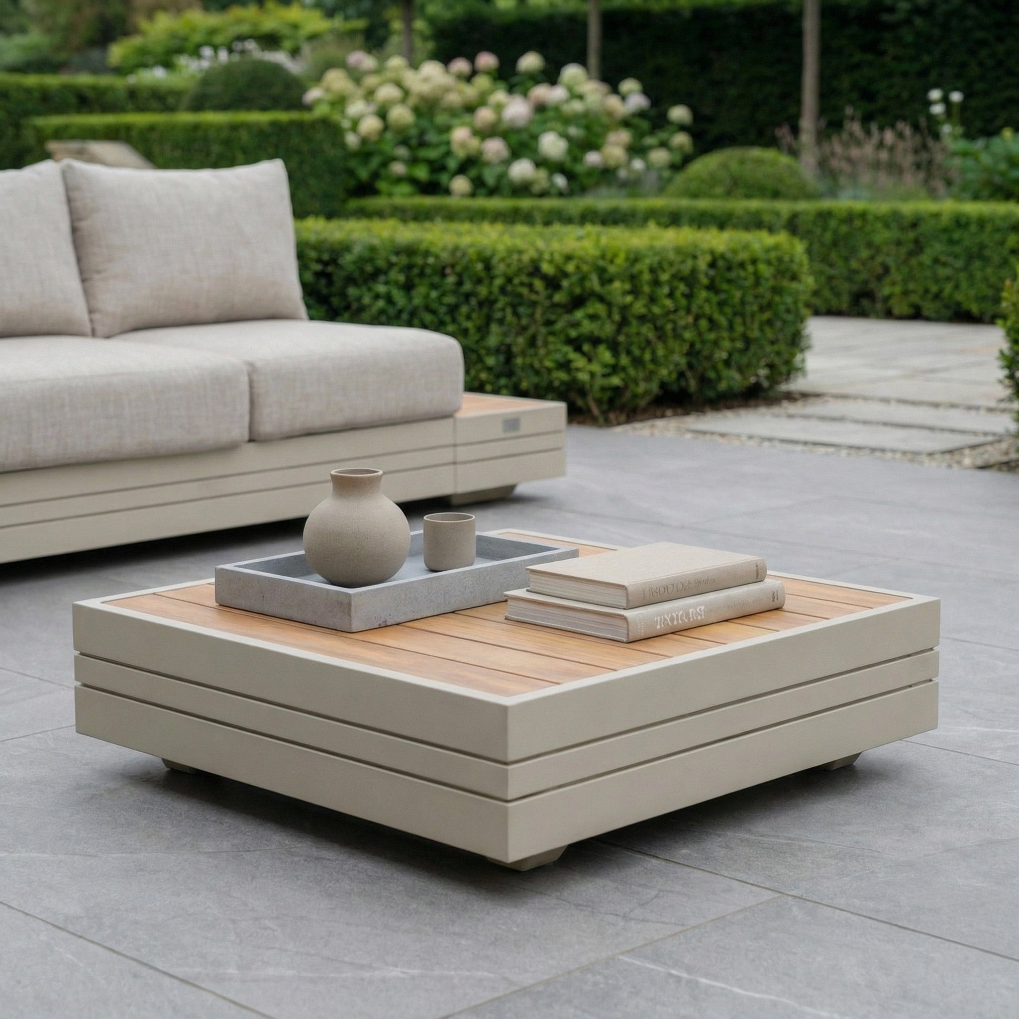 Modern outdoor patio with beige sofa, Panama Teak Coffee Table in Latte, vases, books, and neatly trimmed garden hedges.