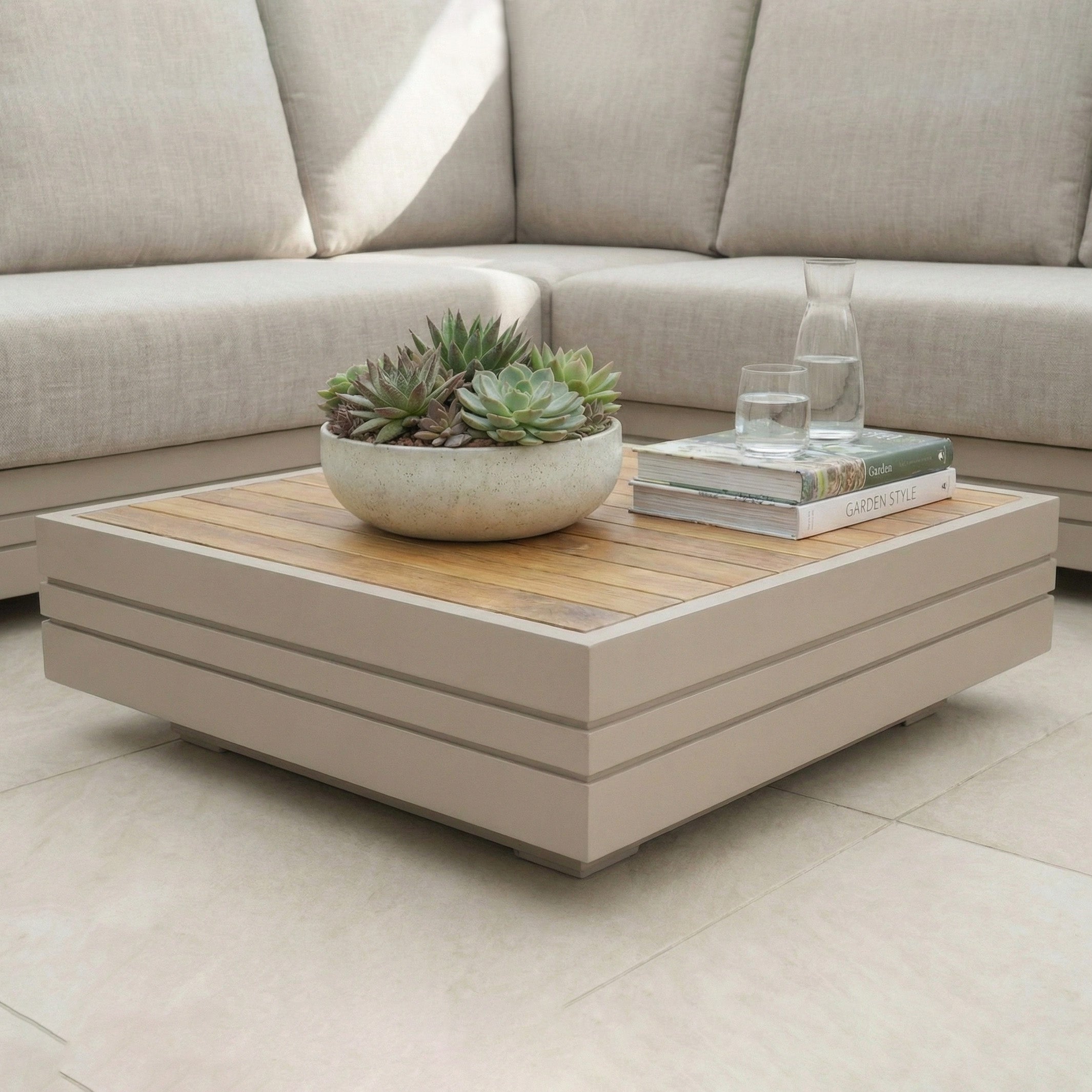 The Panama Teak Coffee Table in Latte, styled with a planter, books, a carafe, and glasses, adds modern flair to your living room.
