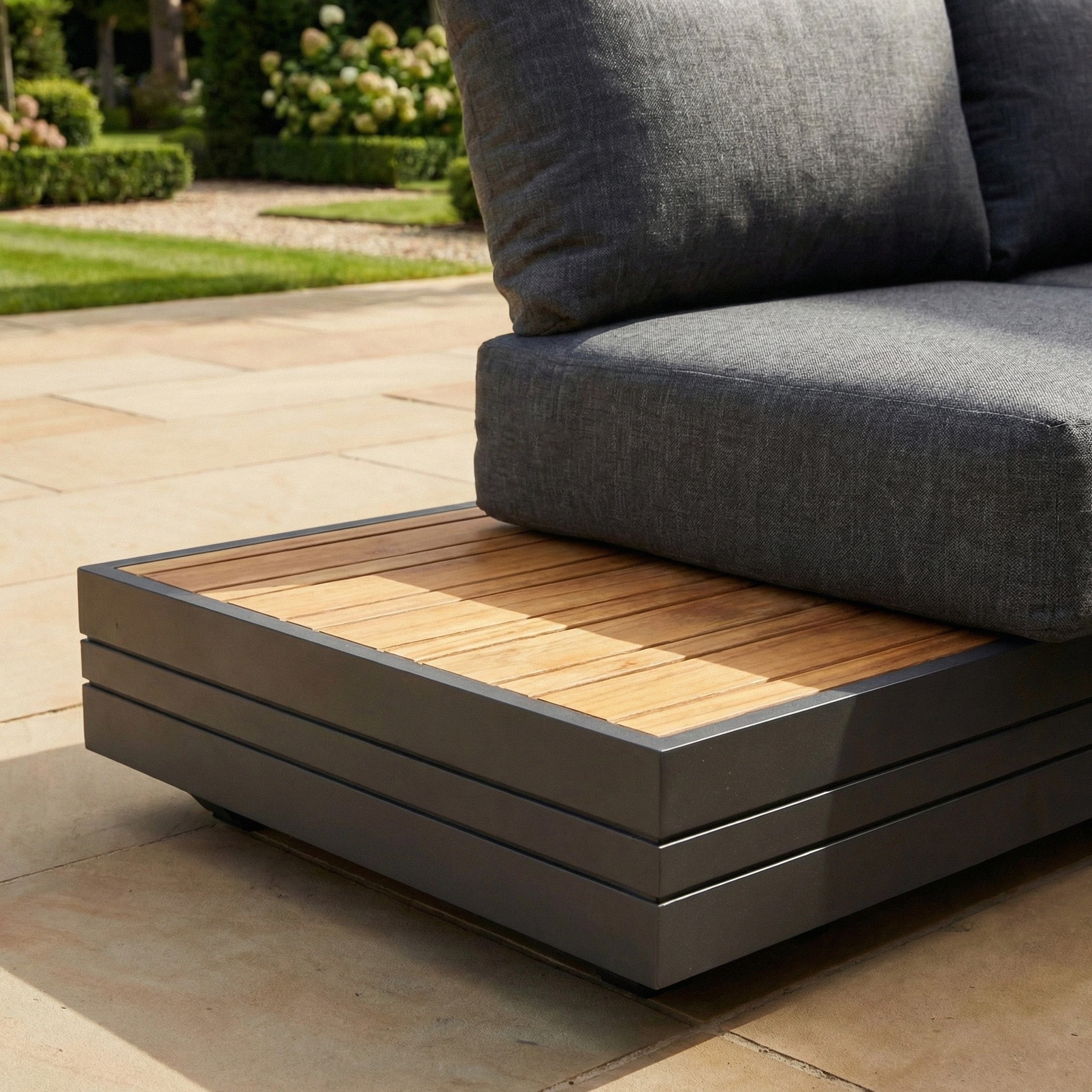 A modern outdoor sofa with gray cushions and a wooden platform base, paired with the Panama Teak Side Table, set on a stone patio.