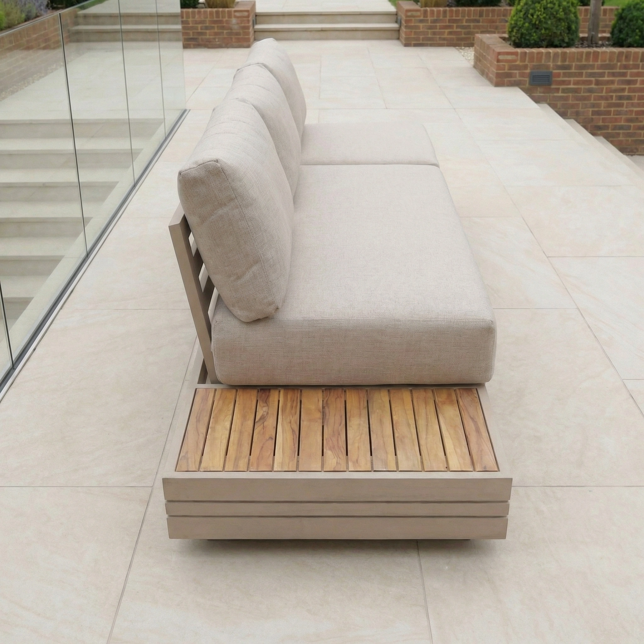 Beige outdoor sofa with wooden slats and aluminium frame on a tiled patio, complemented by the Panama Teak Side Table in Latte. Glass railing and stairs in the background complete this stylish outdoor setup.