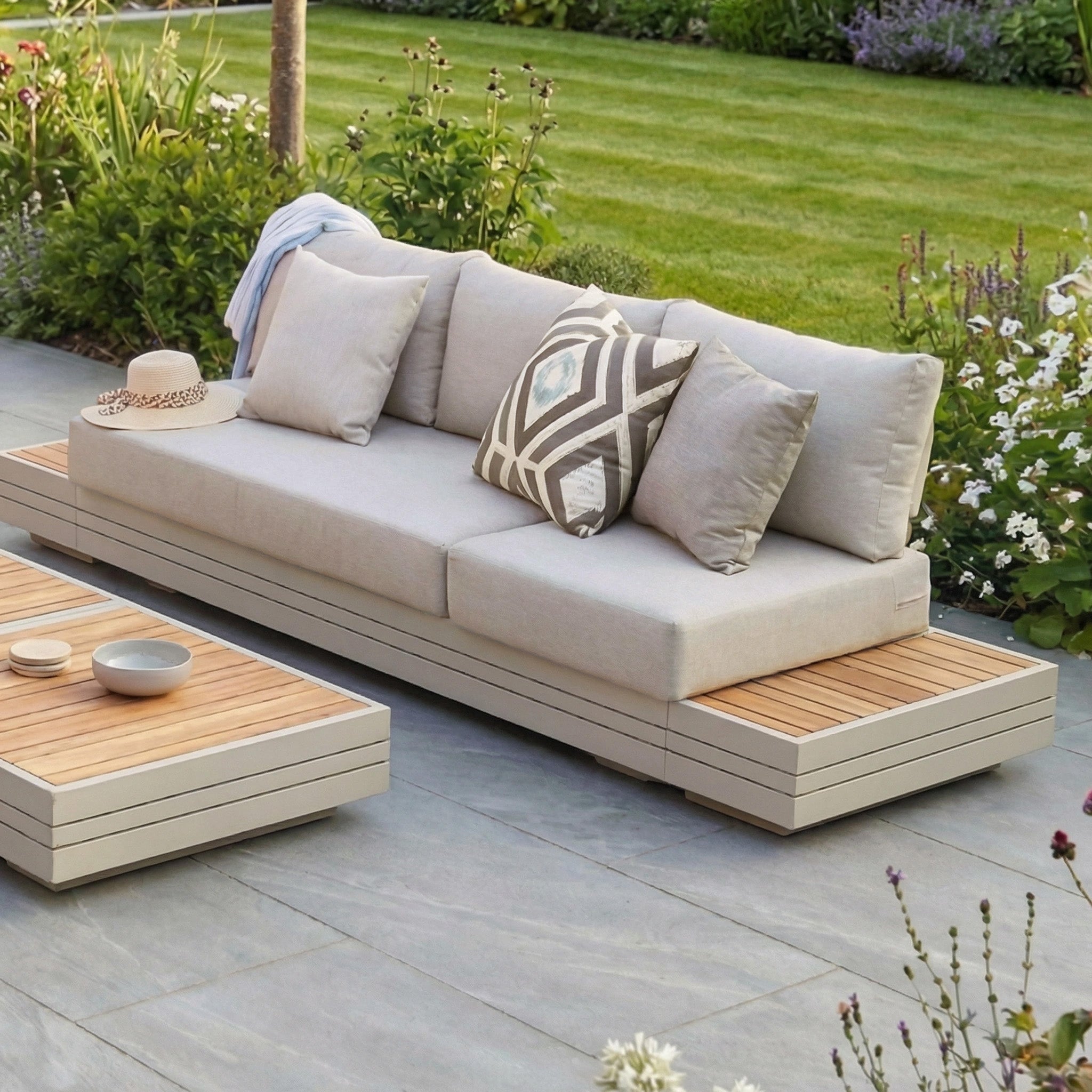 The Panama Teak Side Table in Latte is shown on a patio in a lush garden, next to a modern outdoor sofa with neutral cushions and a hat on the armrest.