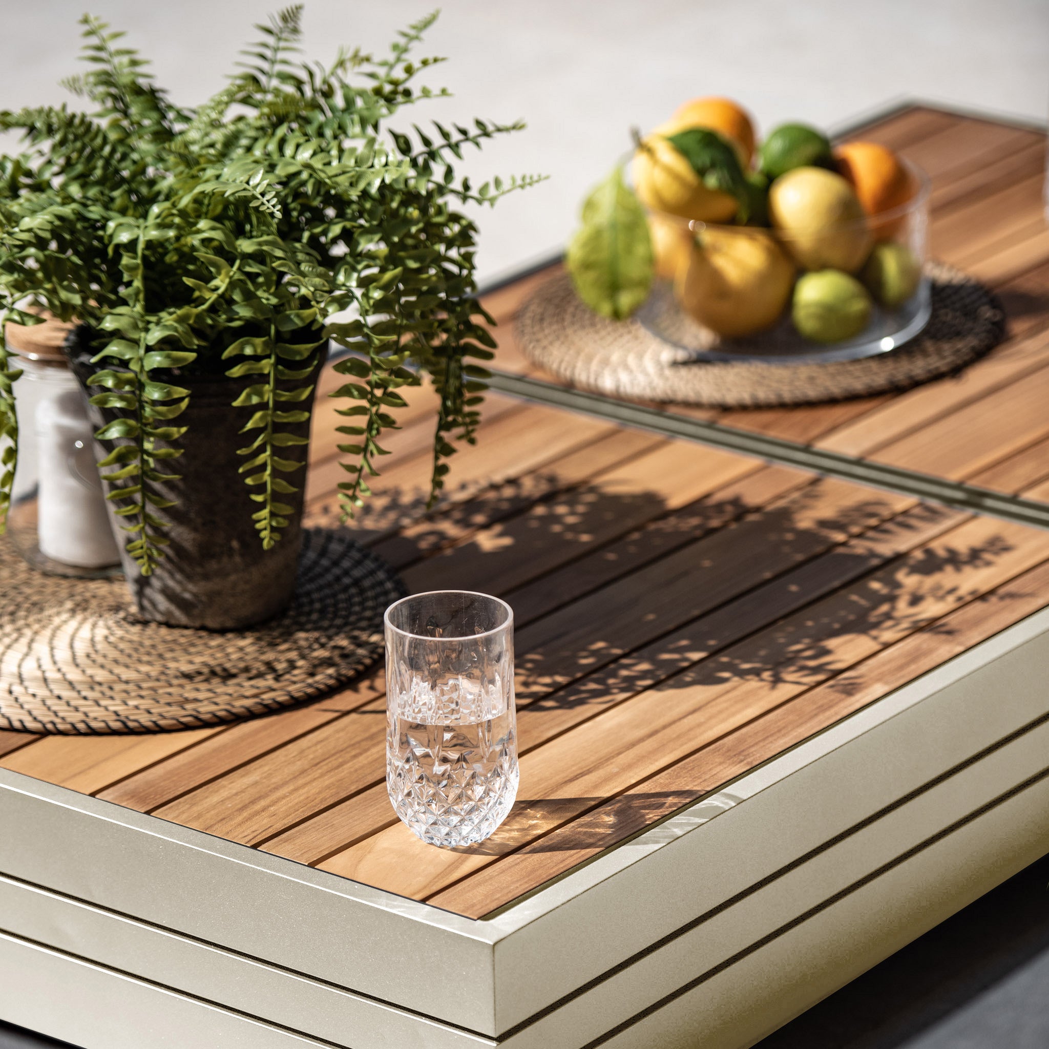 Close-up of the Panama Teak Coffee Table in Latte, featuring a potted fern, a bowl of citrus fruits, and a crystal glass on its elegant wooden surface.