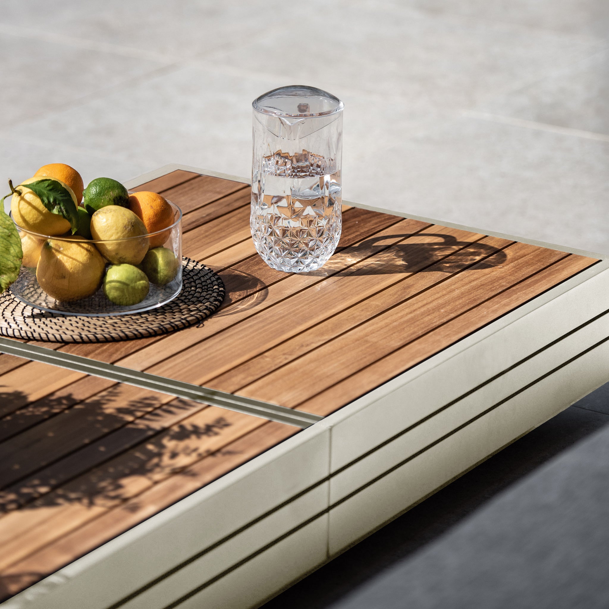 A Panama Teak Coffee Table in Latte holds a glass of water and a bowl of assorted citrus fruits, bathed in natural light.