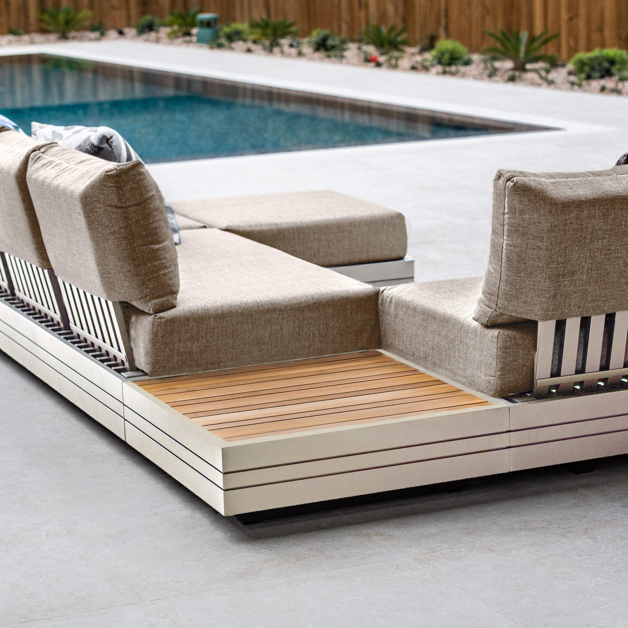 Modern outdoor patio set with beige cushions, wooden accents, and an aluminium frame near a swimming pool with a wooden fence, featuring the luxurious Panama Teak Coffee Table in Latte.