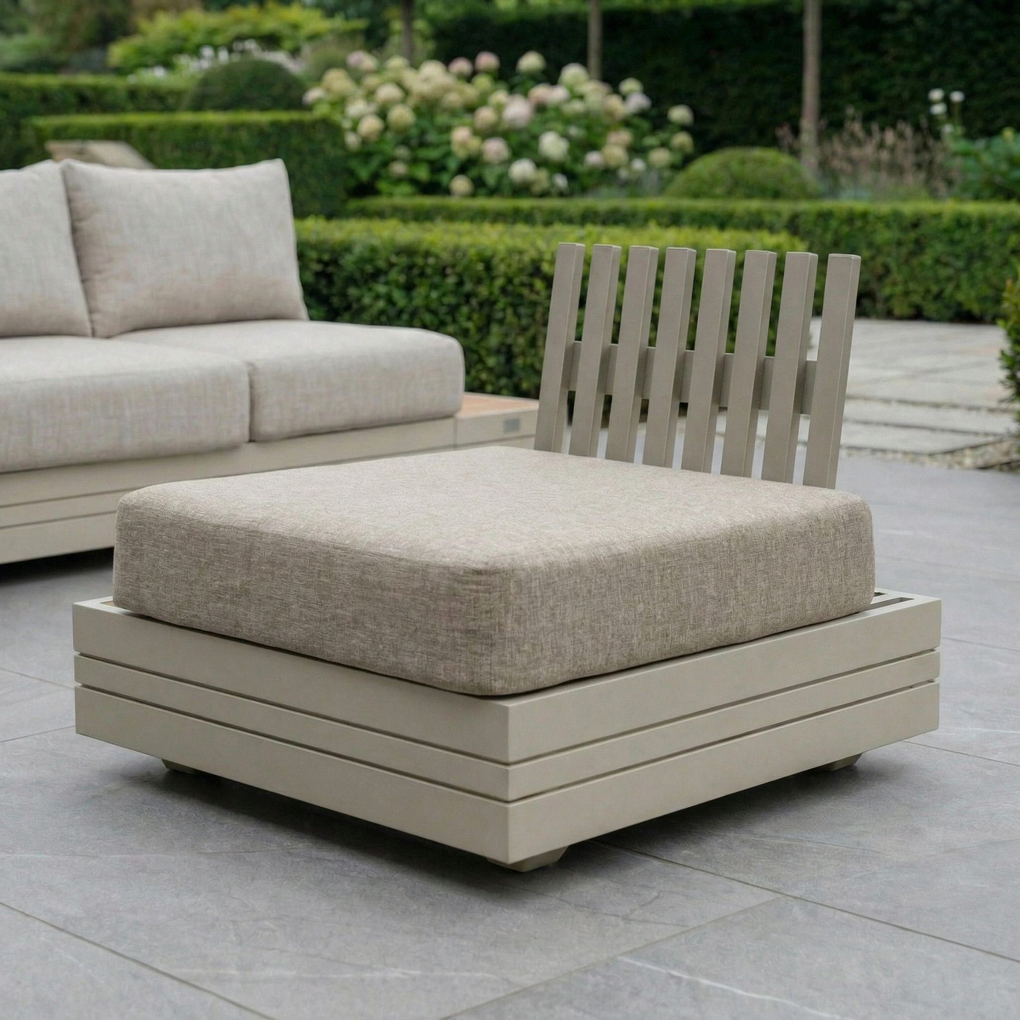 The Panama Corner Unit/Footstool in Latte features a beige cushion and matching footstool, perfect for relaxing on a grey tiled patio overlooking a lush garden.
