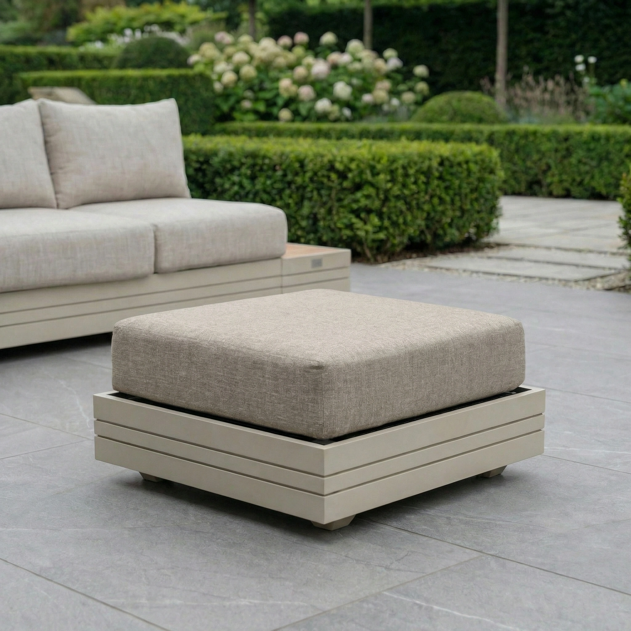 Contemporary outdoor patio with the Panama Corner Unit/Footstool in Latte, featuring beige cushions and set against neatly trimmed green hedges.