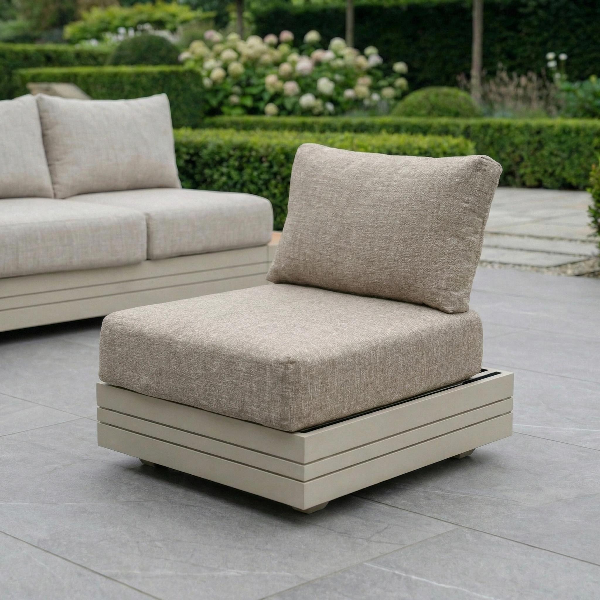 The Panama Corner Unit/Footstool in Latte is shown on a tiled patio with a lounge chair and cushion, set among lush greenery in the background.