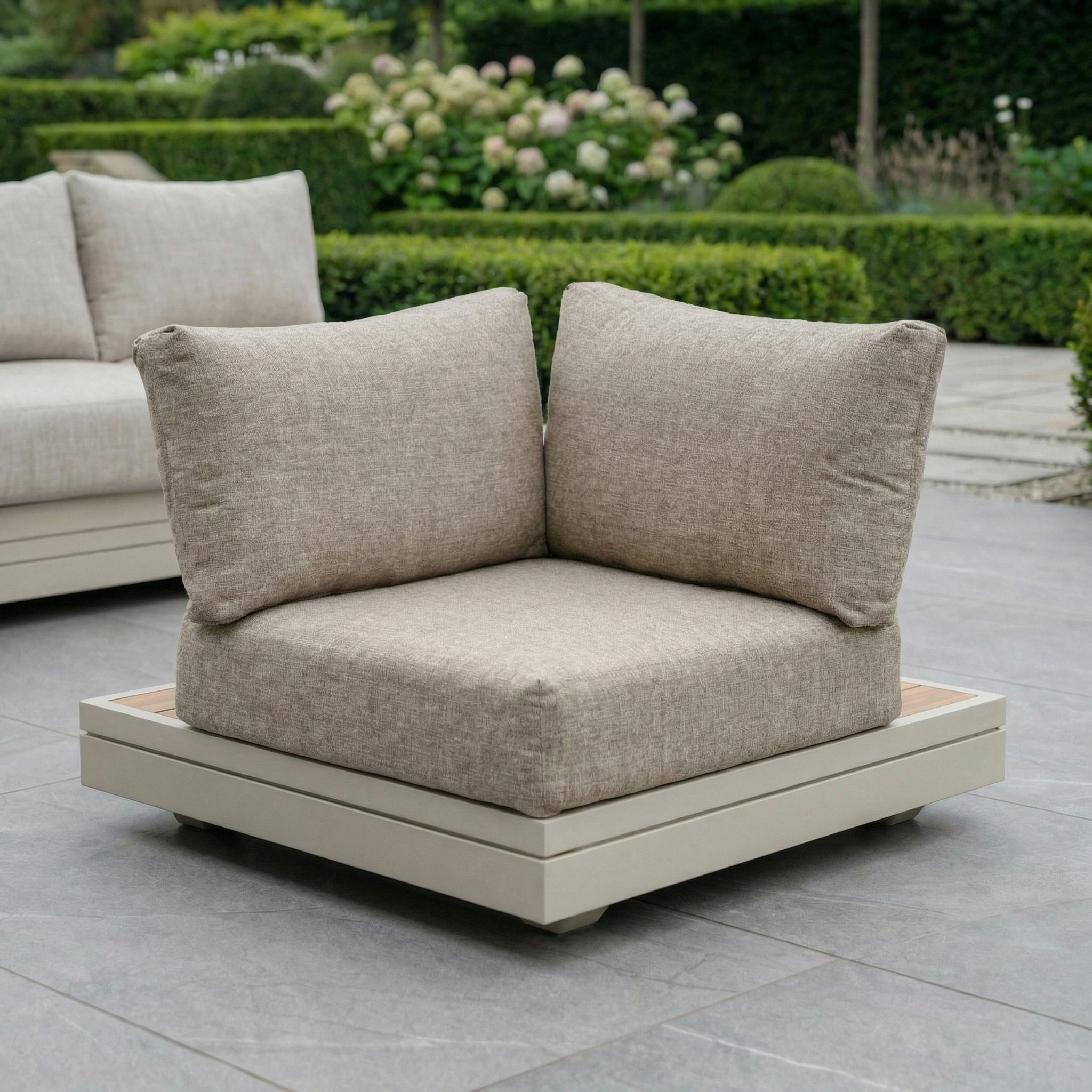 Panama Corner Unit/Footstool in Latte with plush cushions sits on a tiled patio, framed by lush garden greenery in the background.