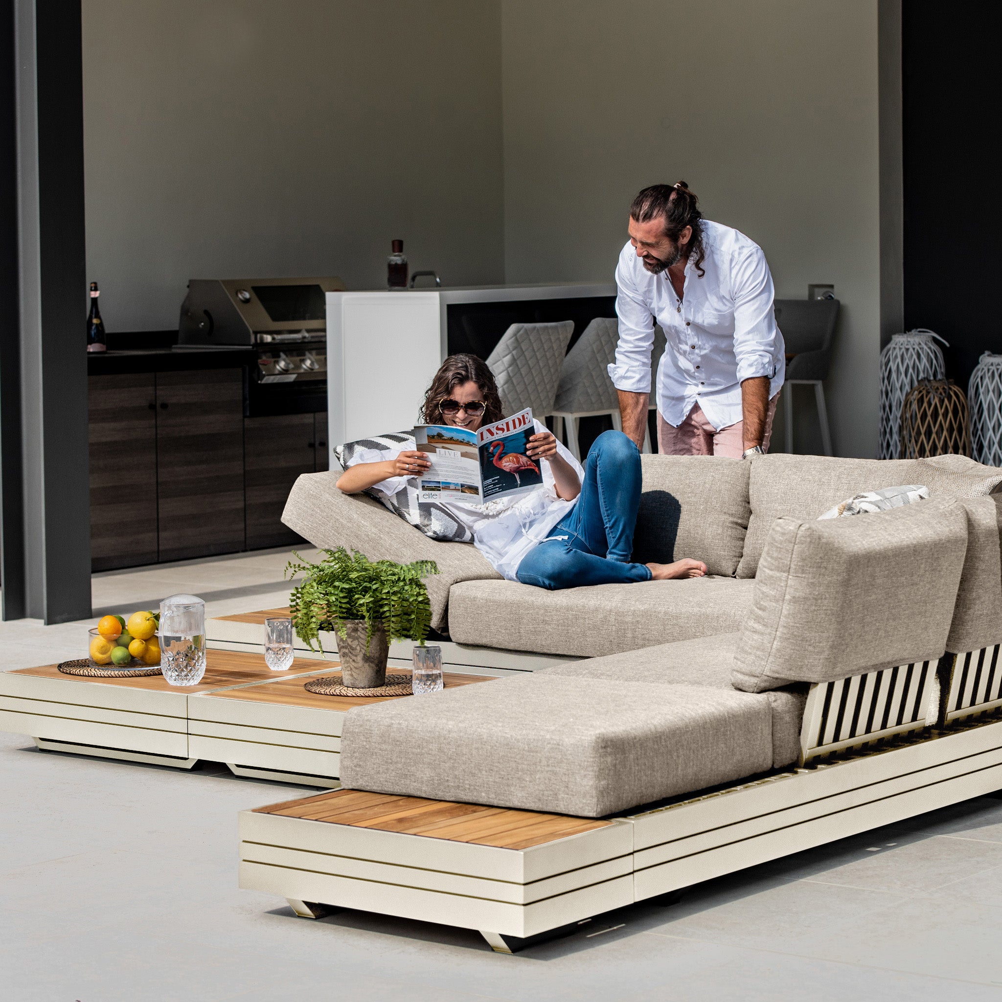 Two people relaxing on an outdoor patio with the Panama Luxury Outdoor Large Corner Group Set in Latte; one is reading a magazine on the sustainable teak lounge chair while the other stands nearby.