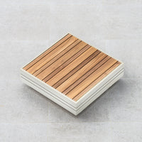 Square wooden deck tile with parallel slats on a light gray tiled floor, perfectly complementing the sleek frame of the Panama Teak Coffee Table in Latte nearby.