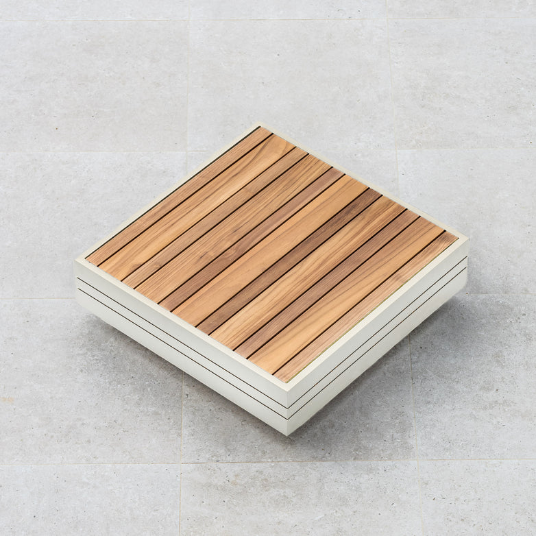 Square wooden deck tile with parallel slats on a light gray tiled floor, perfectly complementing the sleek frame of the Panama Teak Coffee Table in Latte nearby.