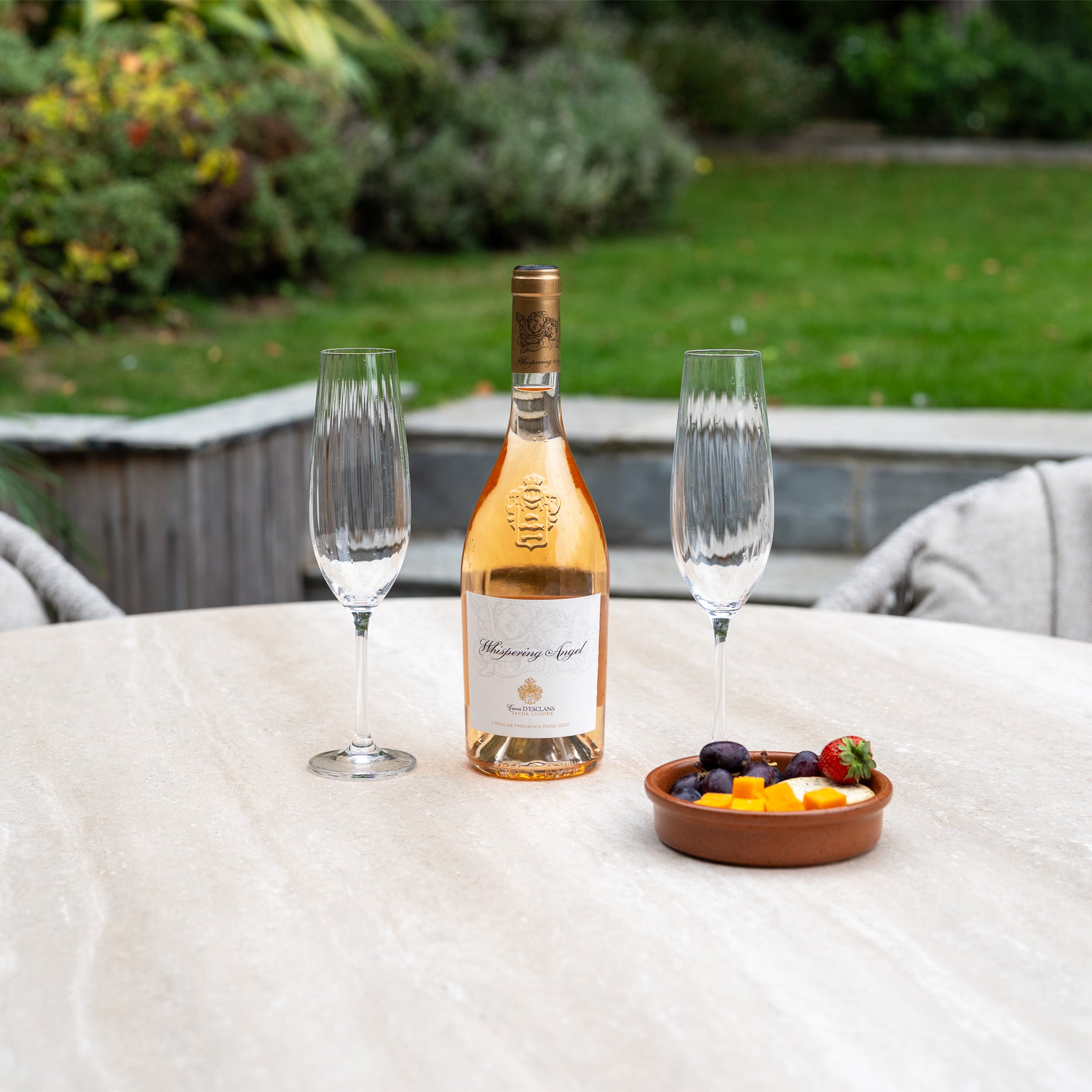 A bottle of rosé, two empty glasses, and fruit sit atop the Pello 4 Seat Round Dining Set with Albatross Table in Fawn—perfect for sharing outdoor moments with your dining furniture.