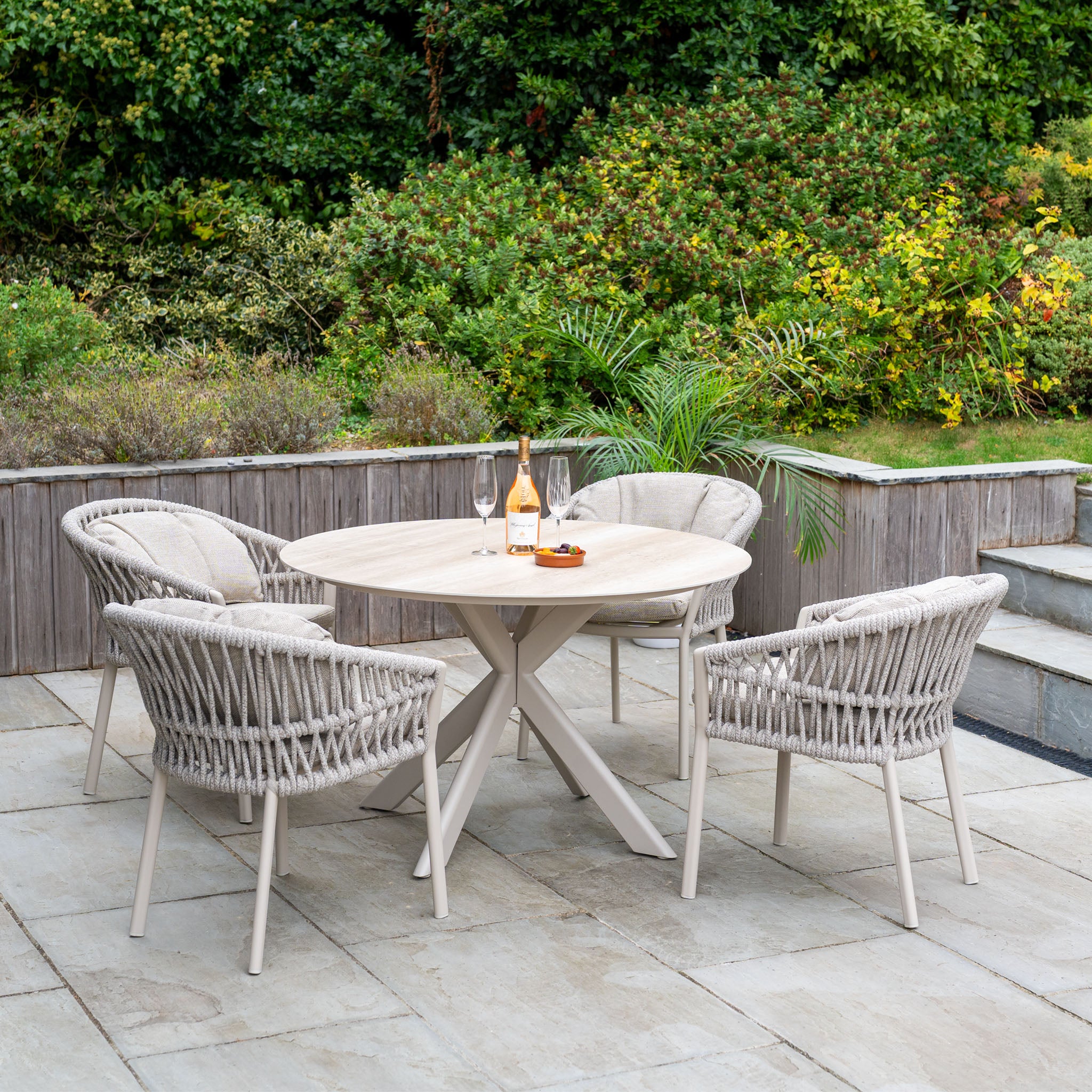 The Pello 4 Seat Round Dining Set with Albatross Table in Fawn features a round patio table and four wicker chairs on a stone terrace with wine, glasses, and snacks—ideal outdoor dining furniture for relaxing amidst lush greenery.