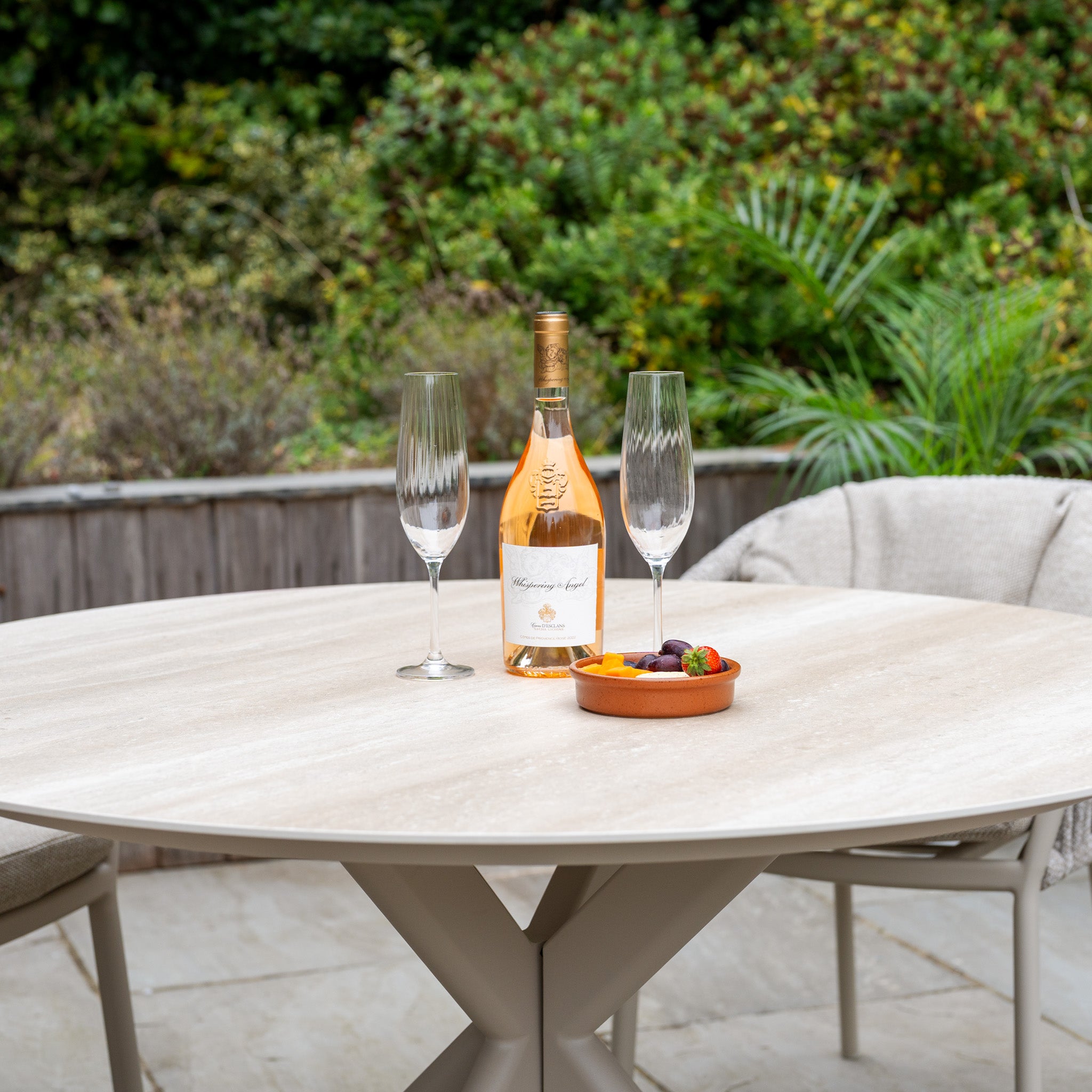 A bottle of rosé, two empty glasses, and a fruit dish adorn the Albatross Table, part of the Pello 4 Seat Round Dining Set with Albatross Table in Fawn—ideal for outdoor dining enthusiasts.