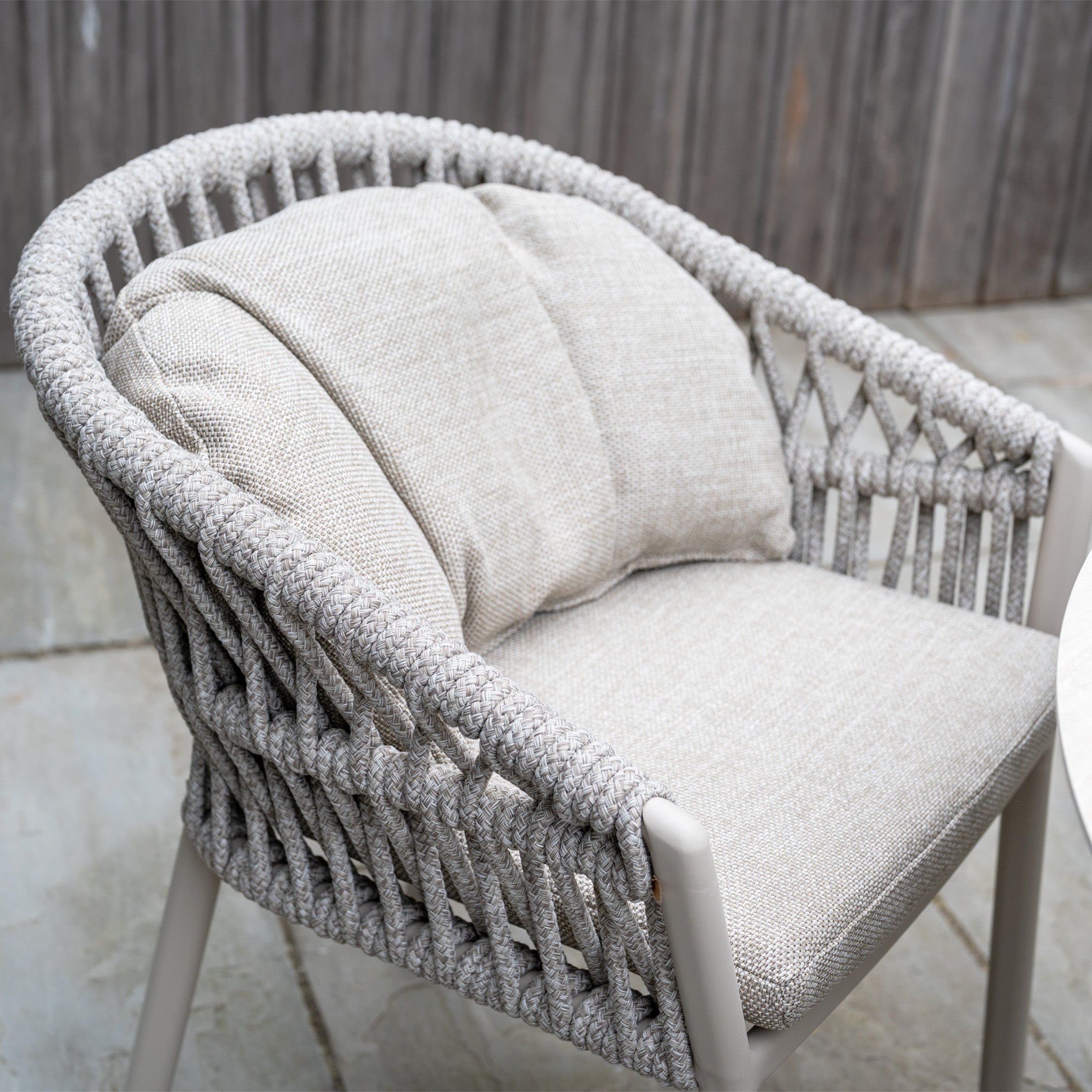 A gray woven outdoor chair with light beige cushions from the Pello 4 Seat Round Dining Set with Albatross Table in Fawn sits on a stone patio by a wooden fence—ideal for stylish outdoor dining.