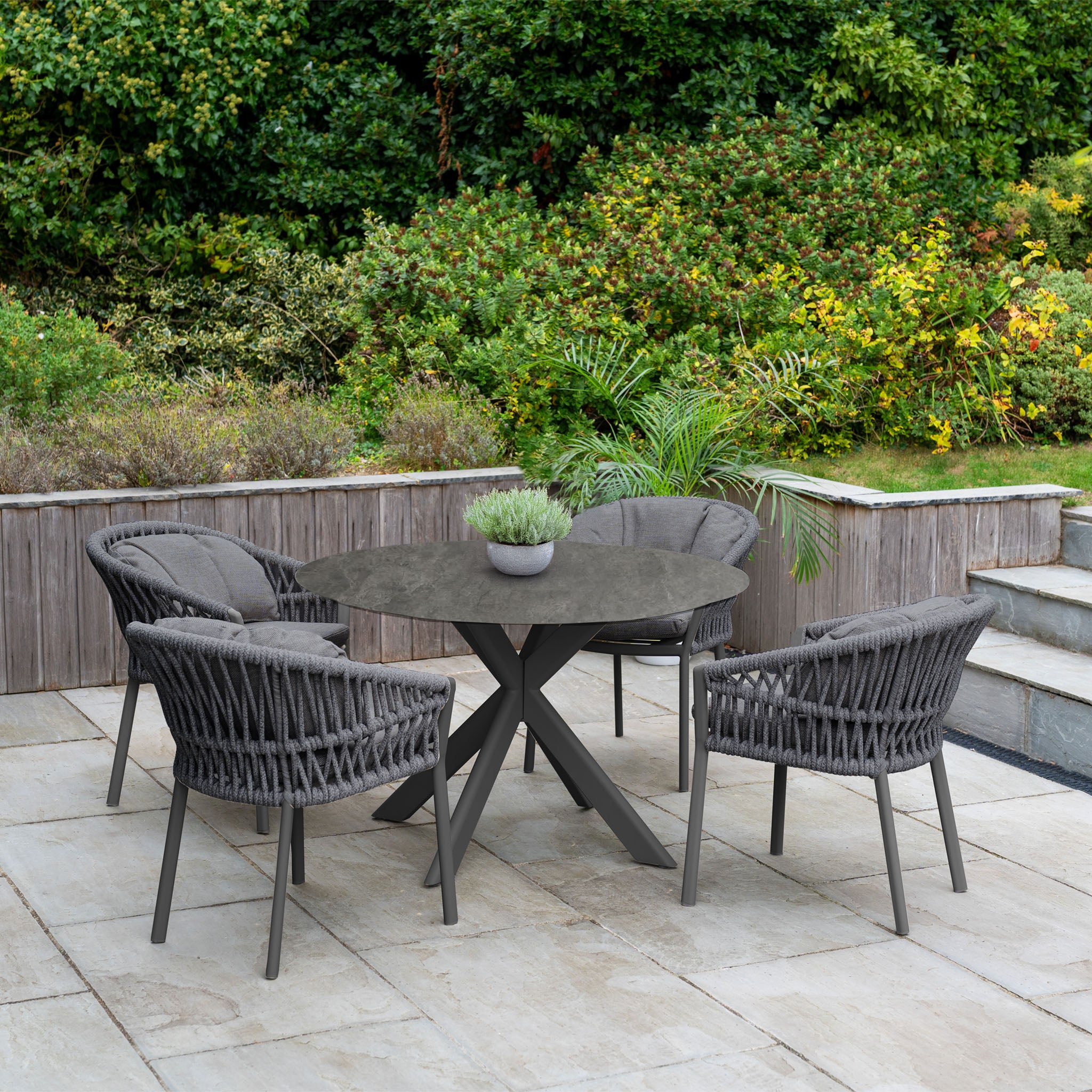 Dine outdoors with the Pello 4 Seat Round Dining Set with Albatross Table in Grey, featuring four wicker chairs and a stylish table, perfect for relaxing on your patio amid lush greenery.
