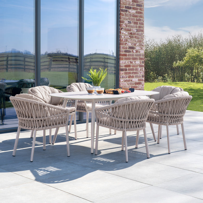 A cozy outdoor patio showcases the Pello 6 Seat Oval Dining Set with a Ceramic Table in Fawn, ergonomic chairs, a modern table, a potted plant, and sunlight reflecting off glass doors.