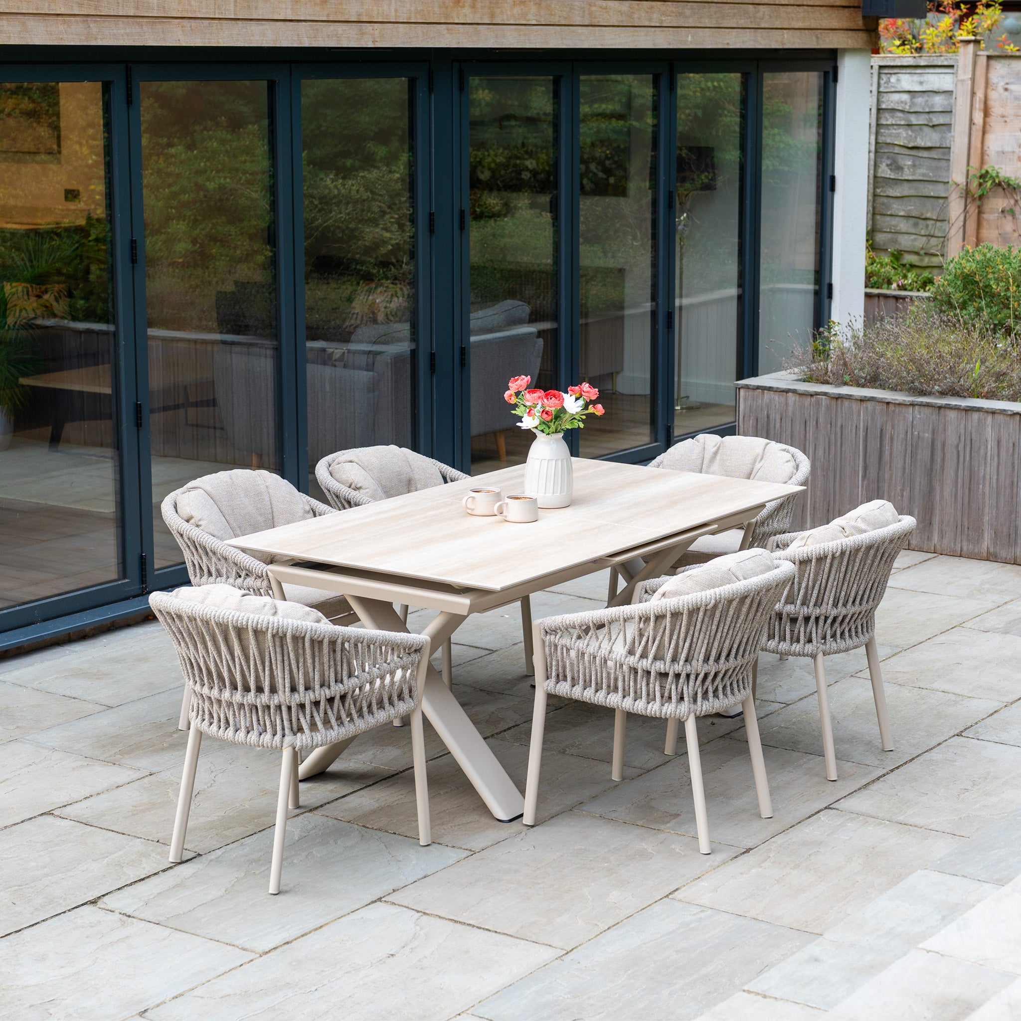 The Pello 6 Seat Dining Set with Albatross Extending Table in Fawn sits on the patio, creating an ideal outdoor dining space.