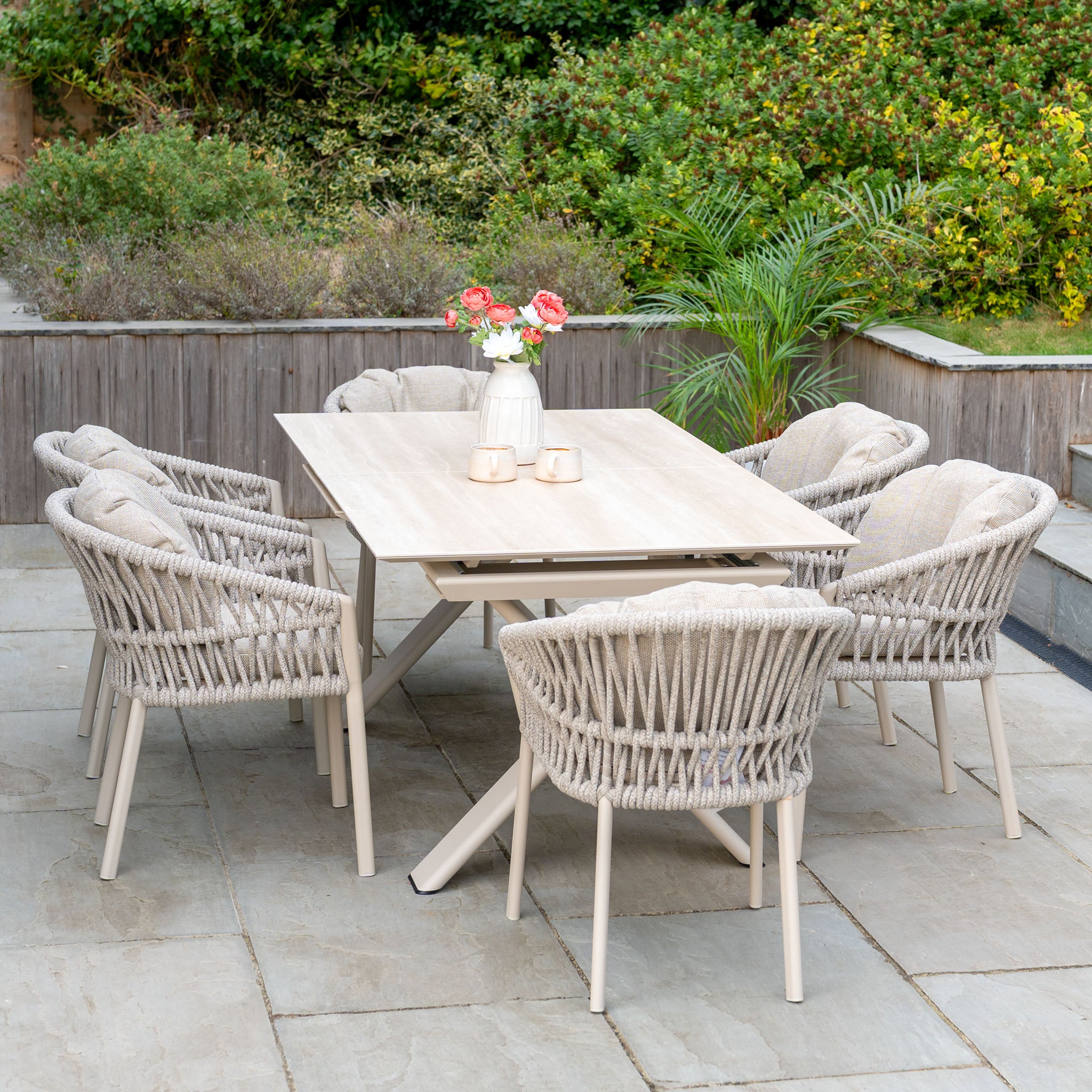 A neutral outdoor dining set, such as the Pello 6 Seat Dining Set with Albatross Extending Table in Fawn, features woven chairs and a light table sitting on a stone patio surrounded by lush greenery.
