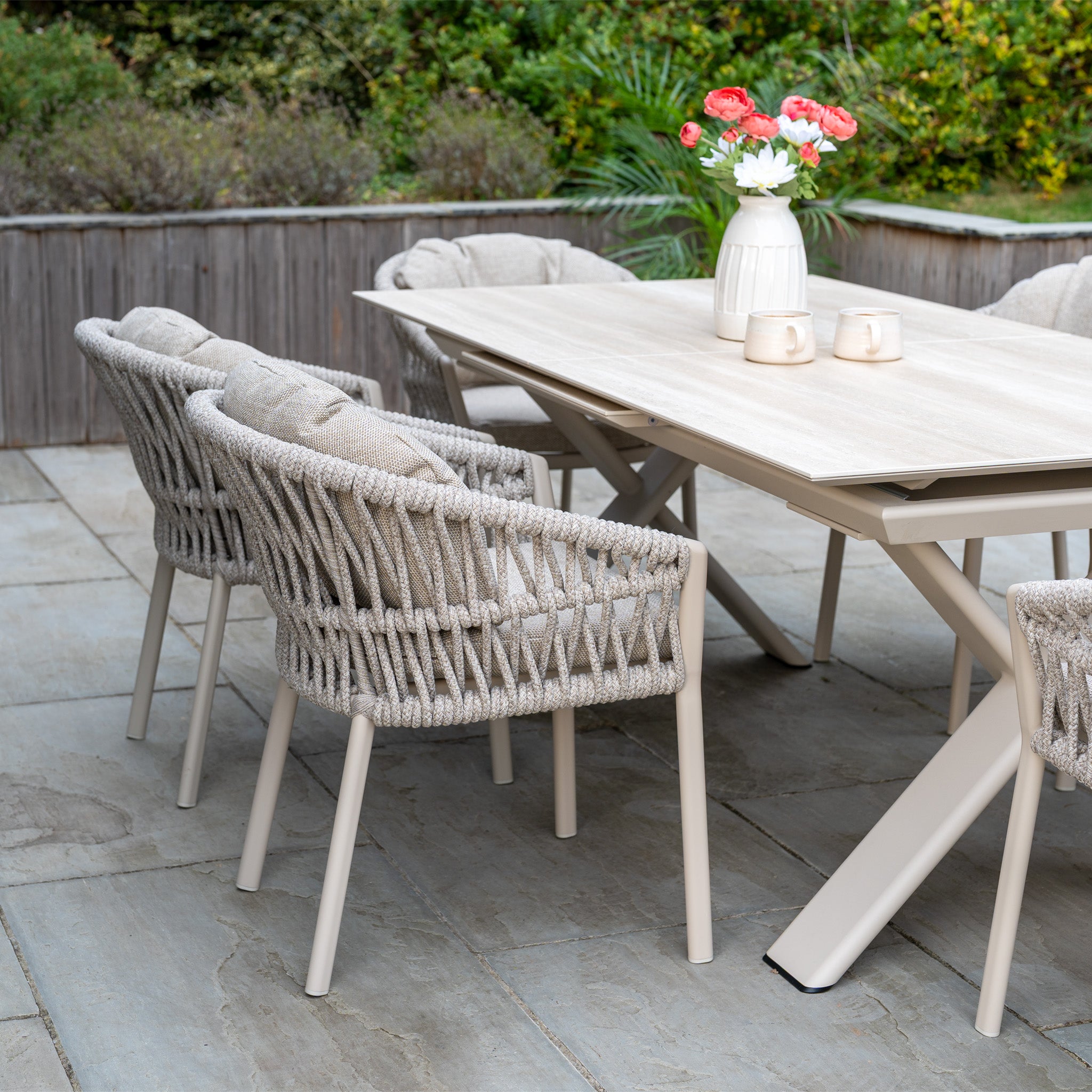 The Pello 6 Seat Dining Set with Albatross Extending Table in Fawn features six seats, perfect for outdoor dining on a stone-tiled patio. Includes woven chairs, a vase of flowers, and two mugs.